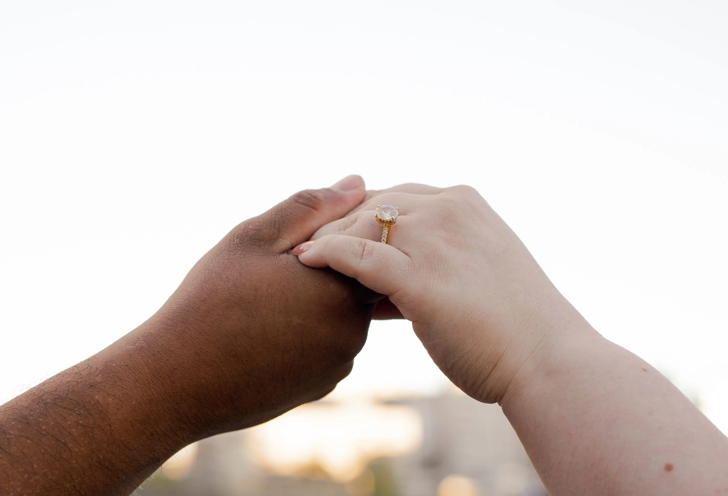 Two hands, one darker and one lighter, holding each other with a large engagement ring on the lighter hand's finger.