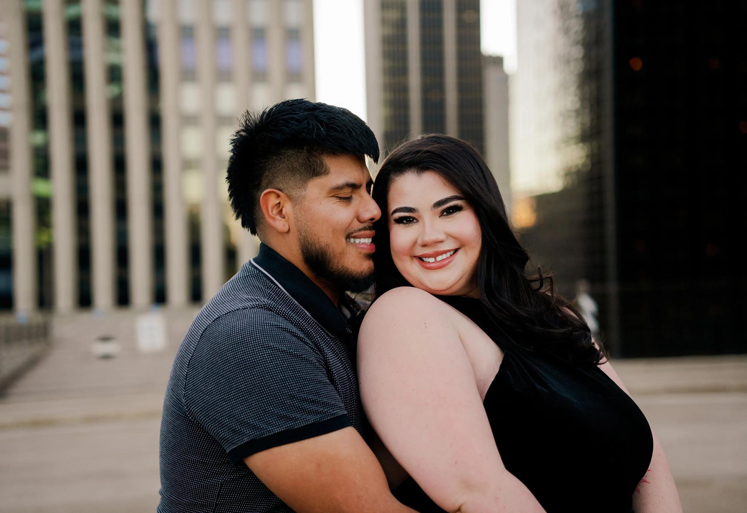 A happy couple stands close together outdoors in an urban setting, with tall buildings in the background. The man has short dark hair, a beard, and is smiling with his eyes closed. The woman has long dark hair, a bright smile, and is wearing a sleeve