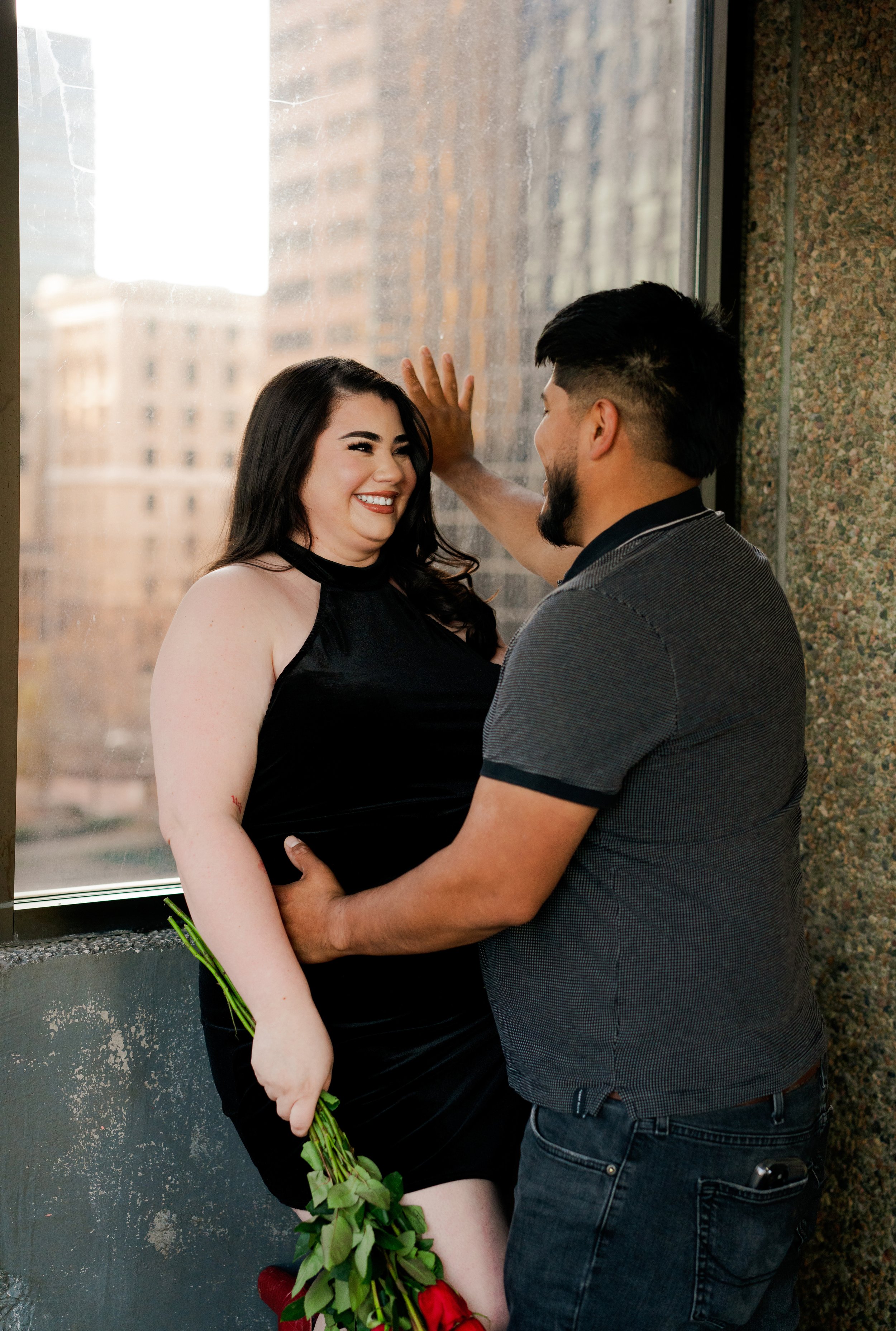 A man and woman are smiling and enjoying a romantic moment by a window, with the city skyline in the background. The woman is holding a bouquet of red roses, and the man is touching the window with one hand while engaging with her.