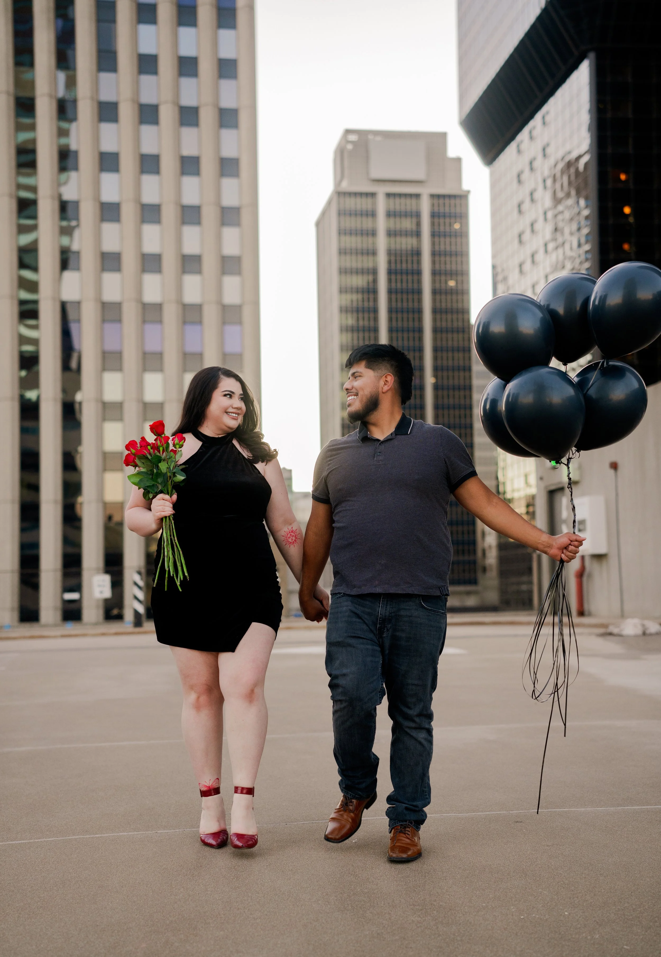 A happy couple holding hands walking in an urban setting with tall buildings, the woman carrying a bouquet of red roses, and the man holding black balloons.