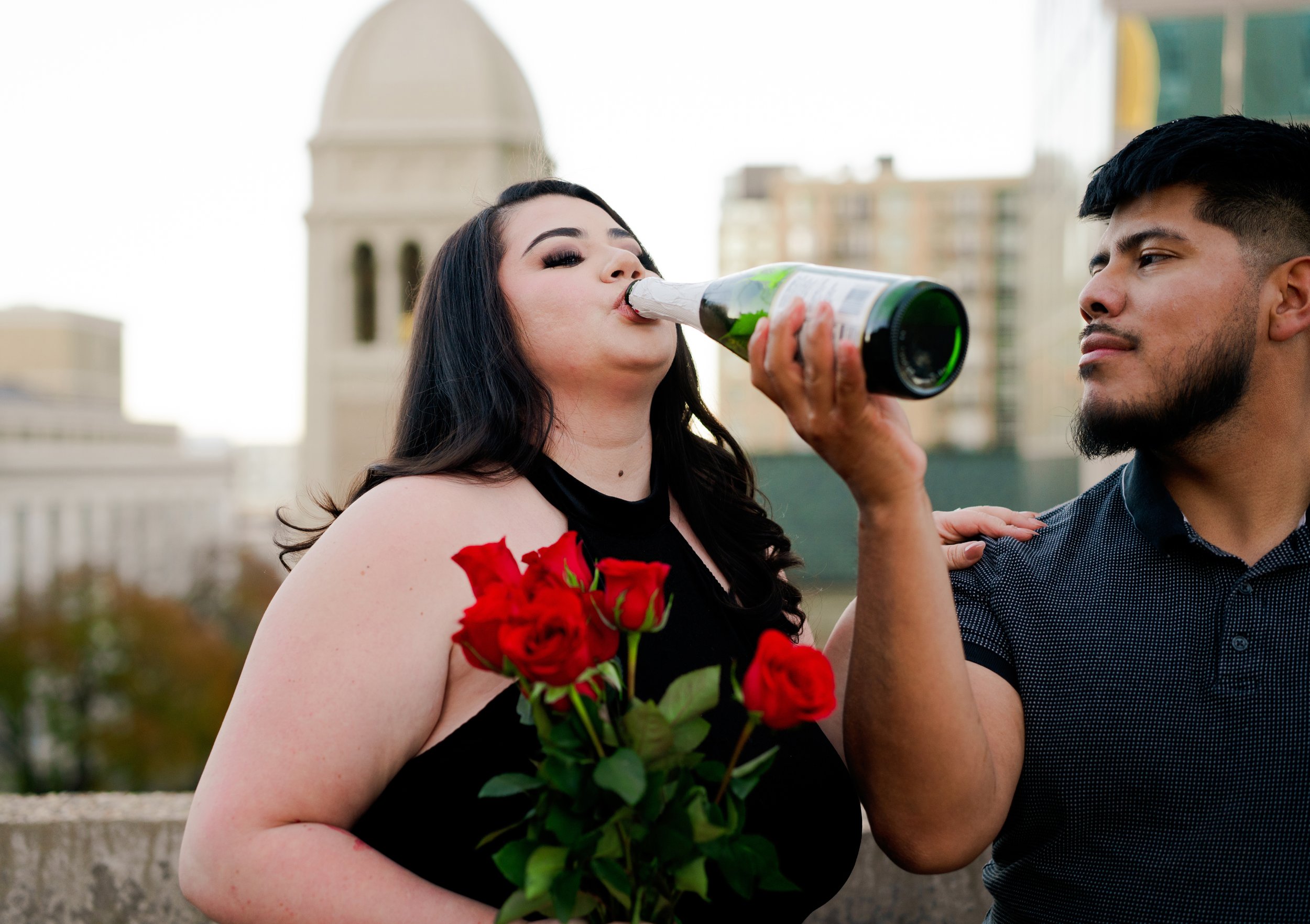 A woman with black hair holding a bouquet of red roses drinks a beer poured from a bottle held by a man with short black hair and a black shirt, in an outdoor urban setting with city buildings in the background.