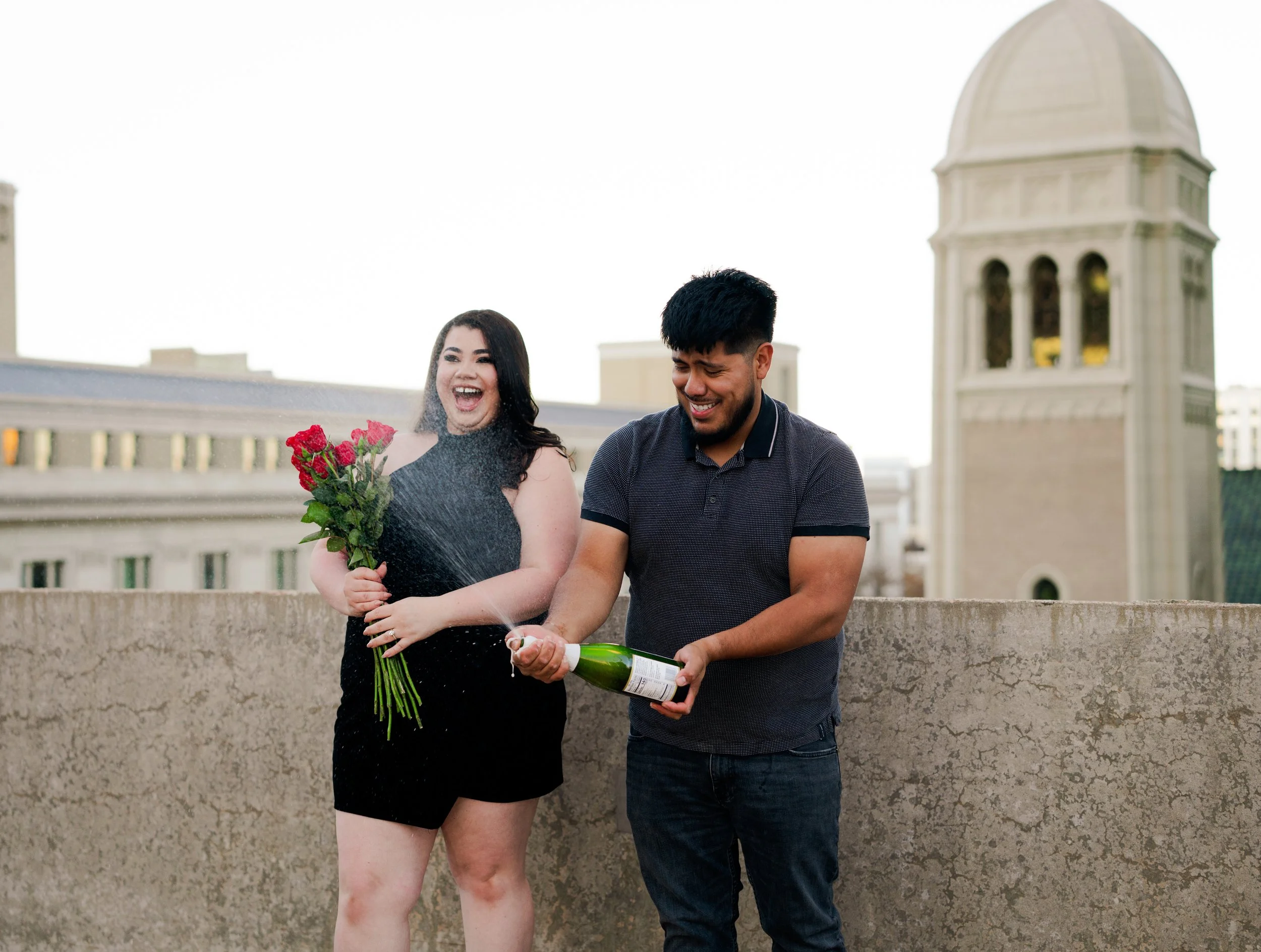 A woman in a black dress holding a bouquet of pink roses and a man in a dark t-shirt opening a bottle of champagne, with spray of champagne, on a rooftop with city buildings and a church tower in the background.