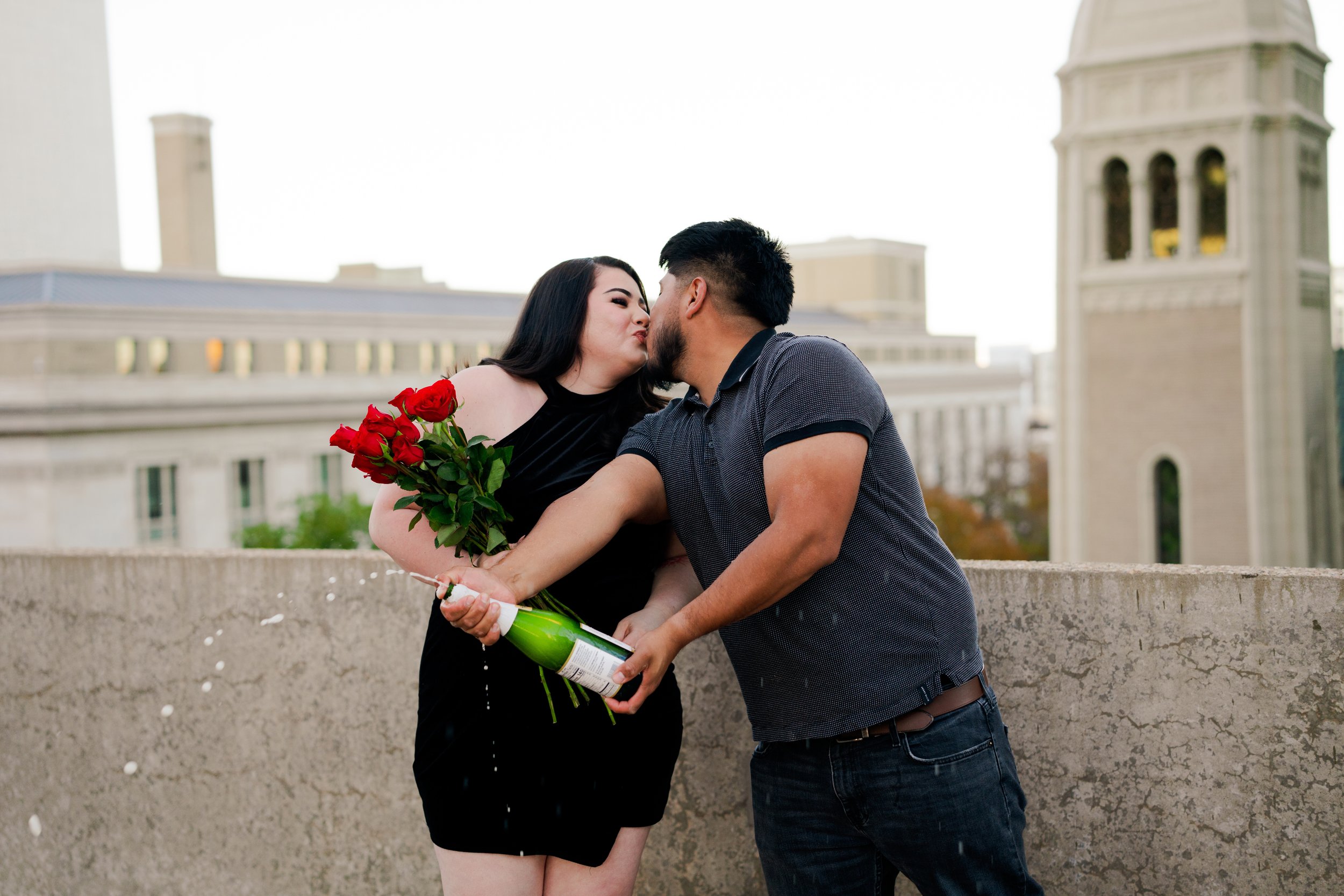 A couple on a rooftop, with the woman holding a bouquet of red roses and a champagne bottle, sharing a kiss during sunset.