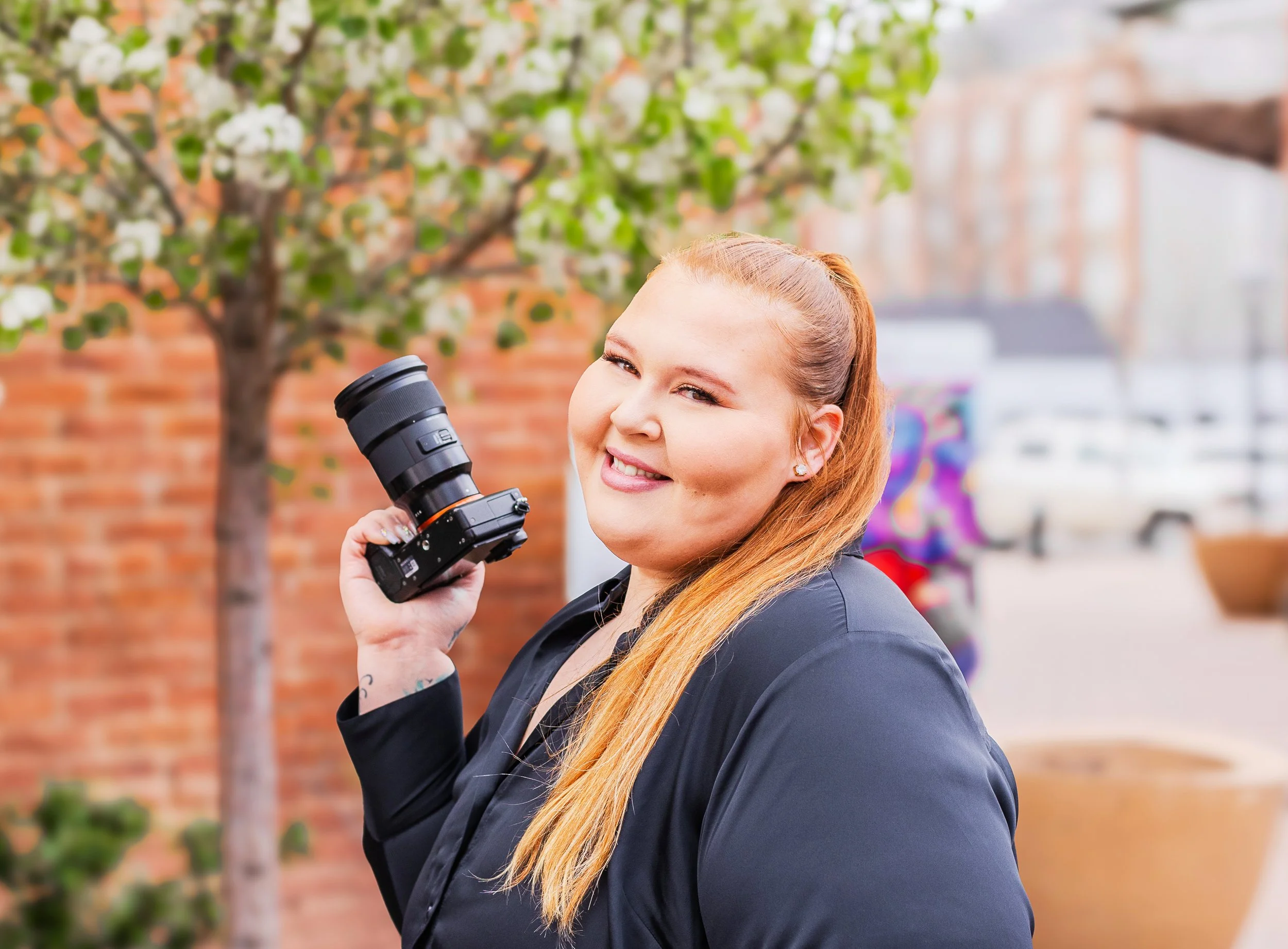 Smiling woman with long red hair holding a camera outdoors in front of a brick wall and a flowering tree.
