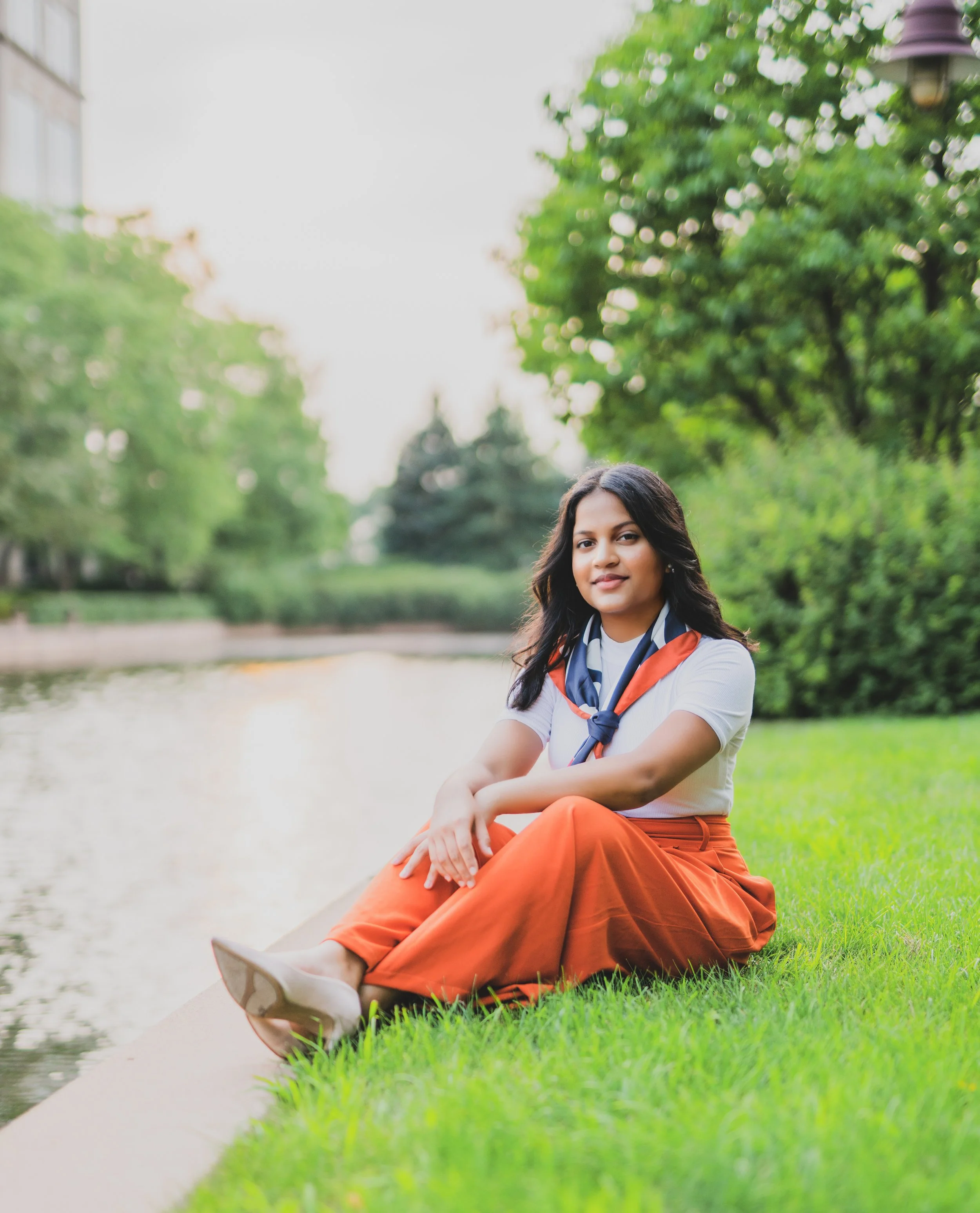 A woman sitting on grass by a pond in a park with green trees in the background
