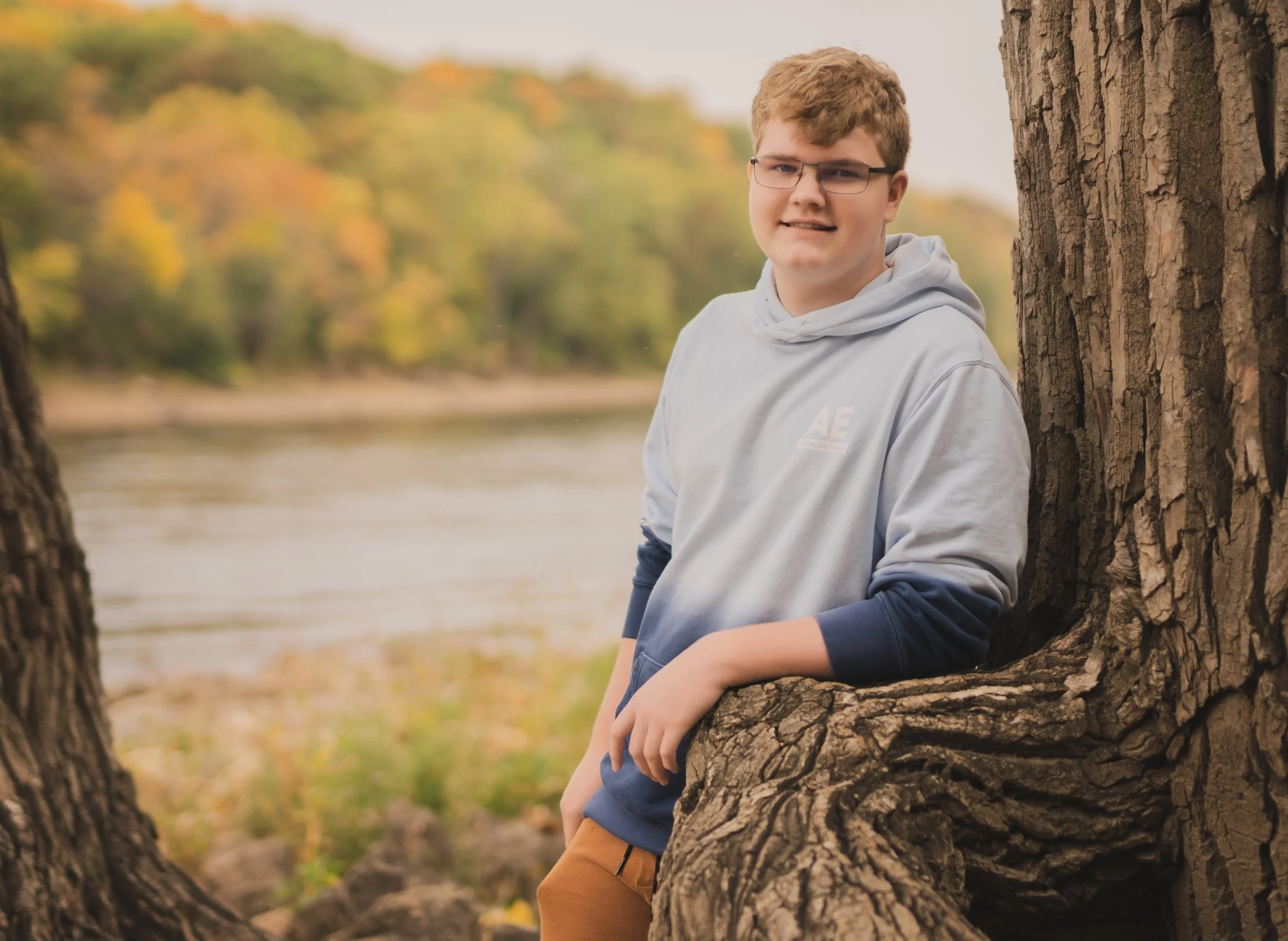 A teenage boy with glasses and curly hair leaning against a large tree trunk near a river, with fall foliage in the background.