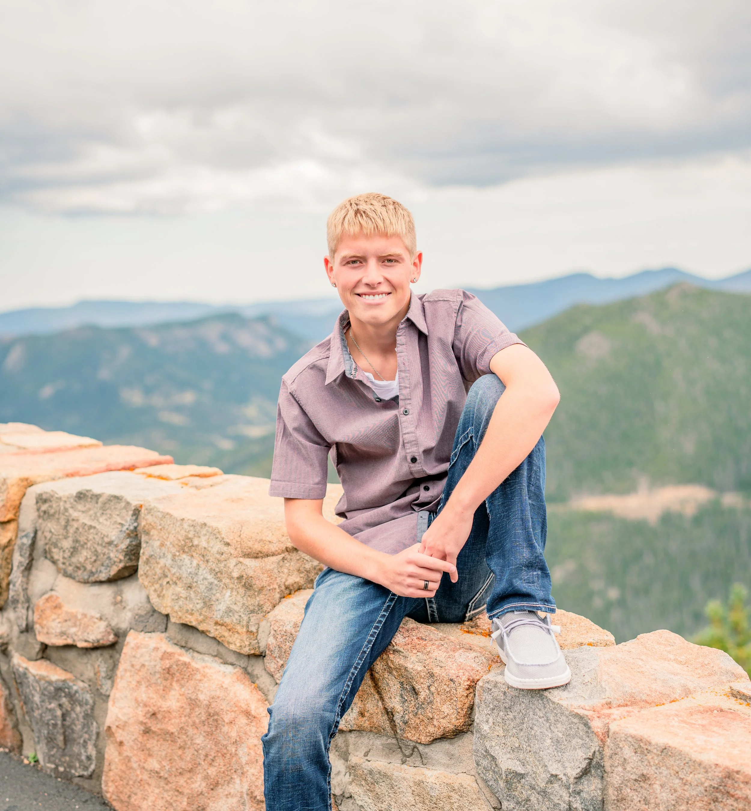 A young person with short blonde hair smiling, sitting on a stone wall with scenic mountains in the background.