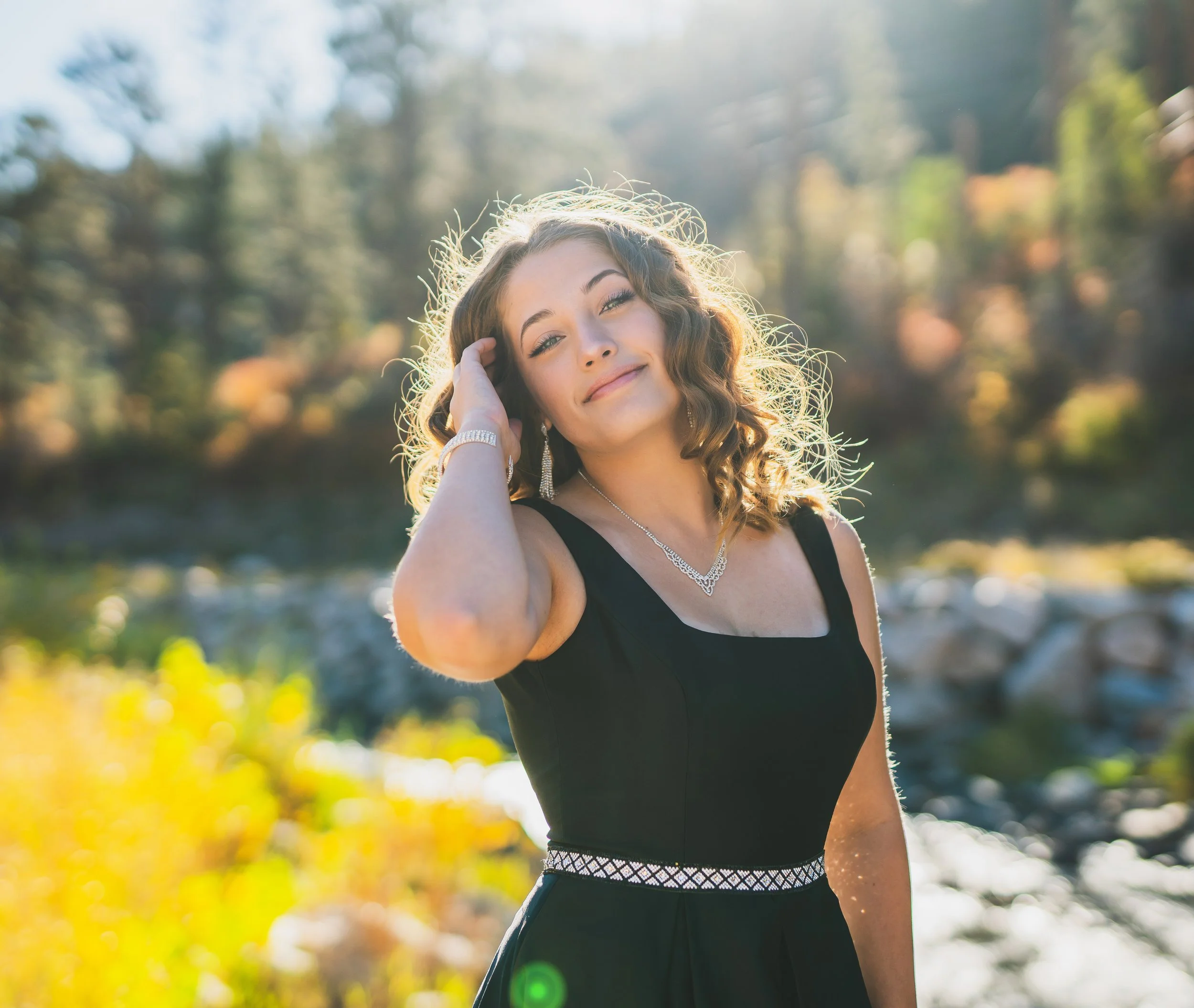 A woman in a black dress with jewelry, outdoors in a sunny, natural setting with trees and rocks in the background.