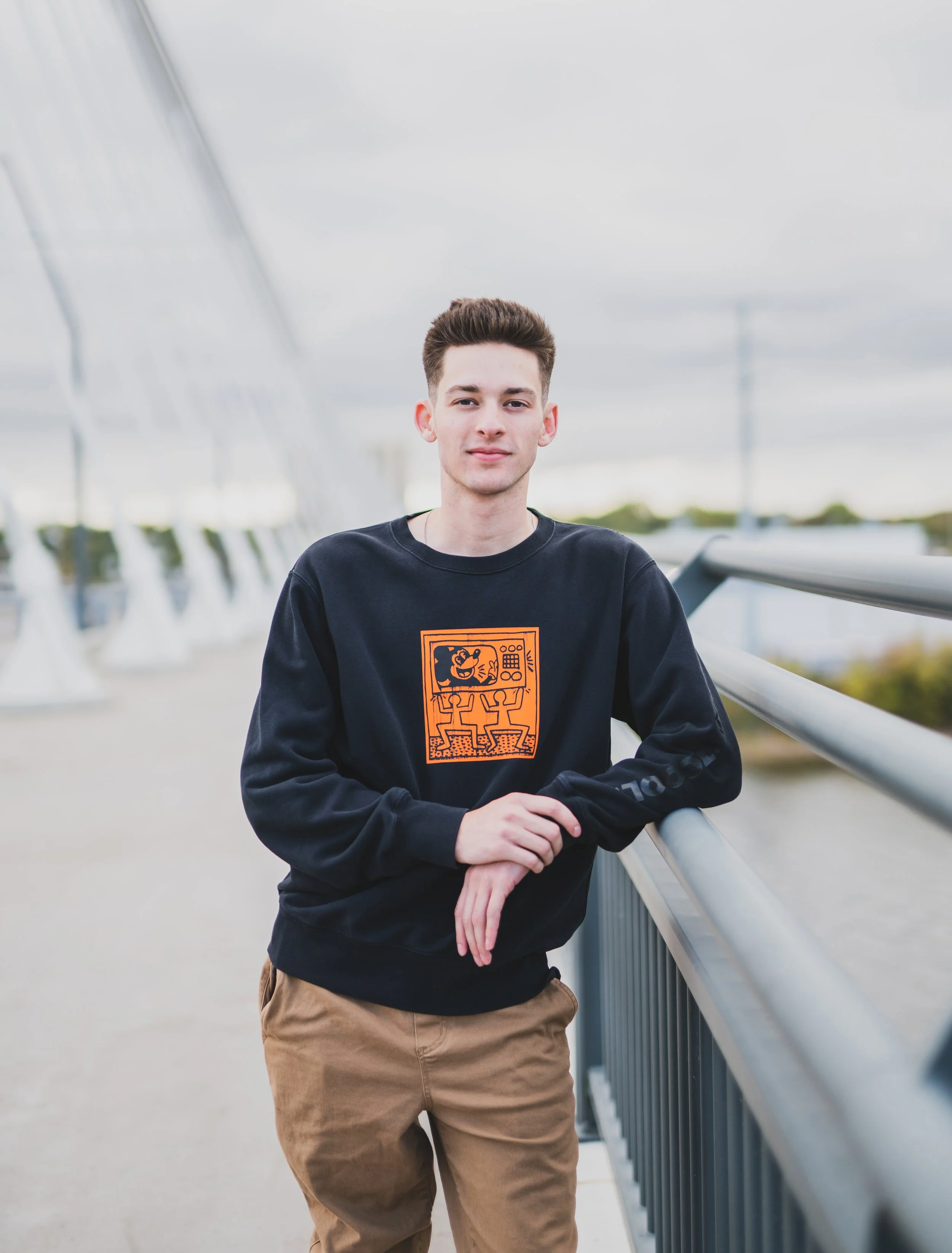 A young man with short brown hair and light skin leaning on a metal railing outdoors, wearing a black sweatshirt with an orange graphic, and tan pants, with a cloudy sky in the background.