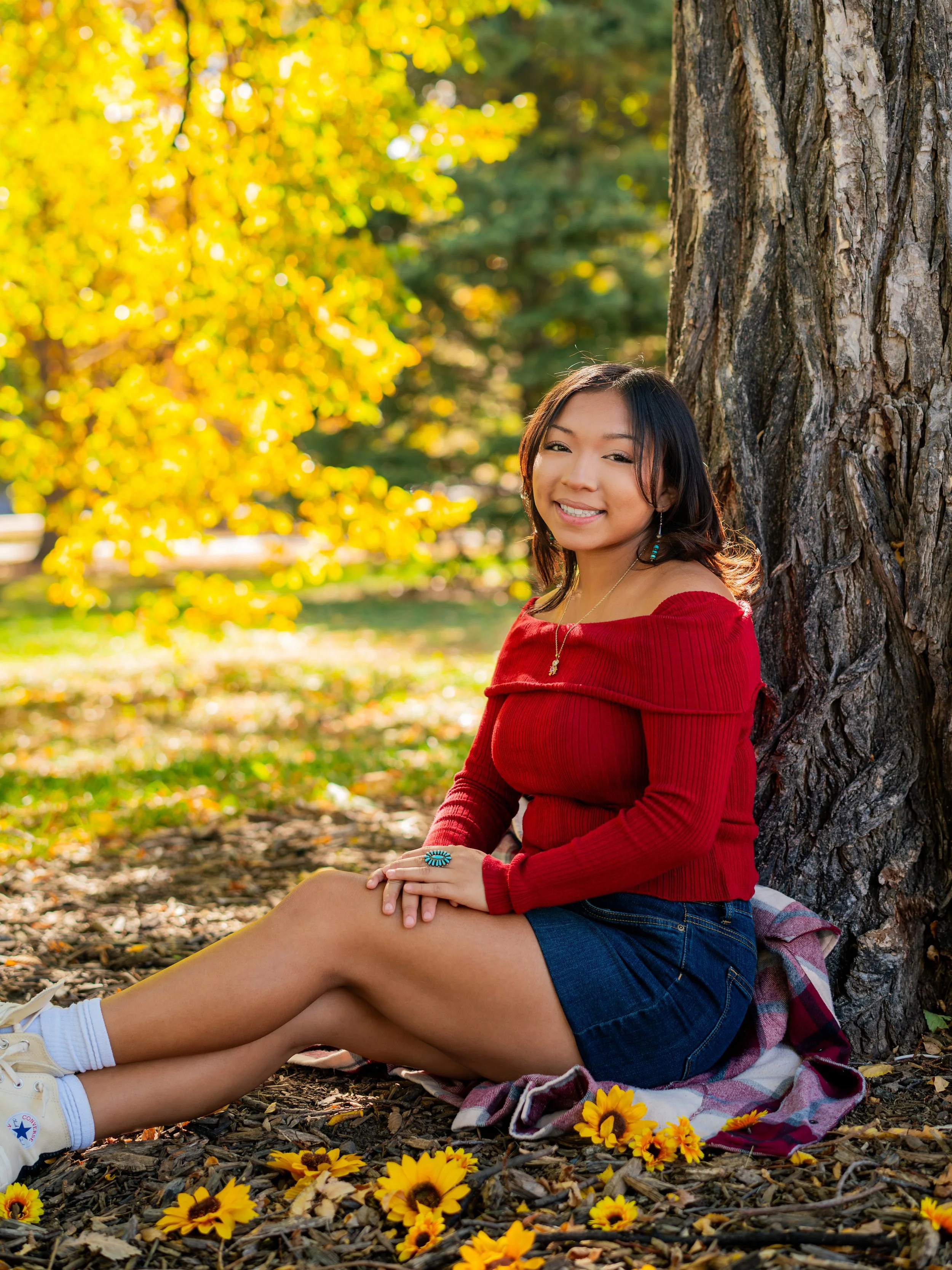 Woman sitting under a tree in a park during autumn, wearing a red sweater and denim shorts, with yellow fall foliage in the background and sunflowers on the ground.
