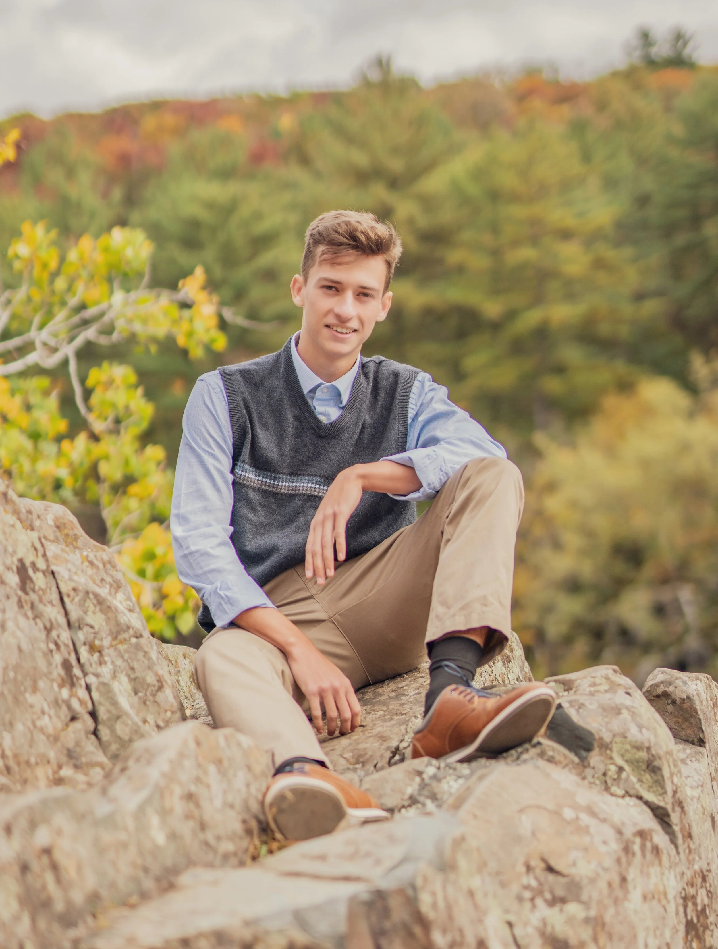 A young man with brown hair, wearing a blue shirt, gray vest, beige pants, and brown shoes, sitting on rocks outdoors with autumn trees in the background.