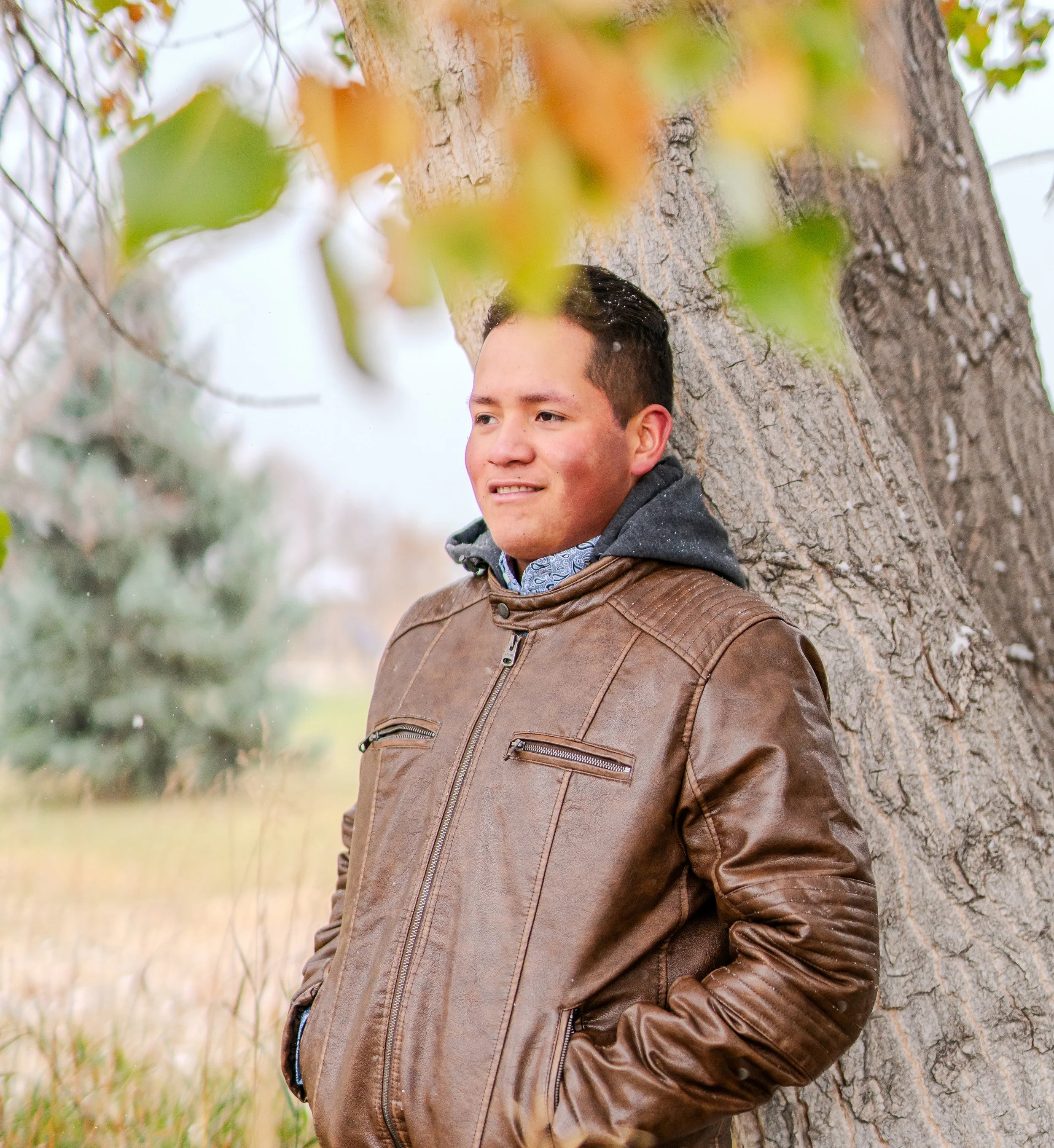 A young man leaning against a tree outdoors during autumn, wearing a brown leather jacket and a gray hoodie, with a slight smile on his face.