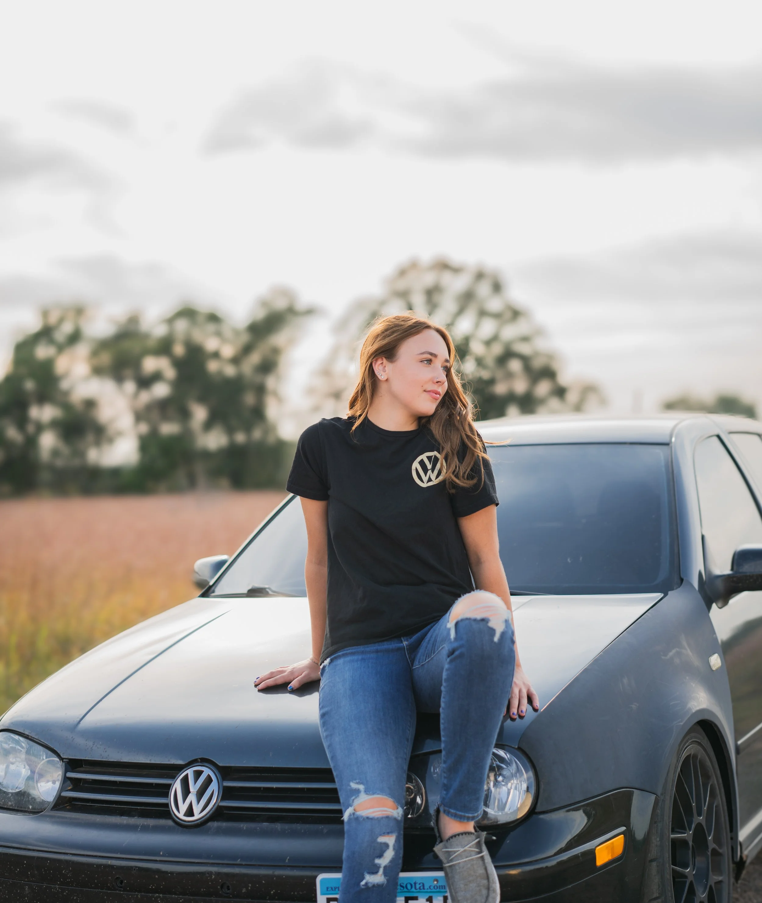 A young woman with long brown hair sitting on the hood of a black Volkswagen car in an open field, wearing a black t-shirt and ripped jeans, looking to the side with a contemplative expression.