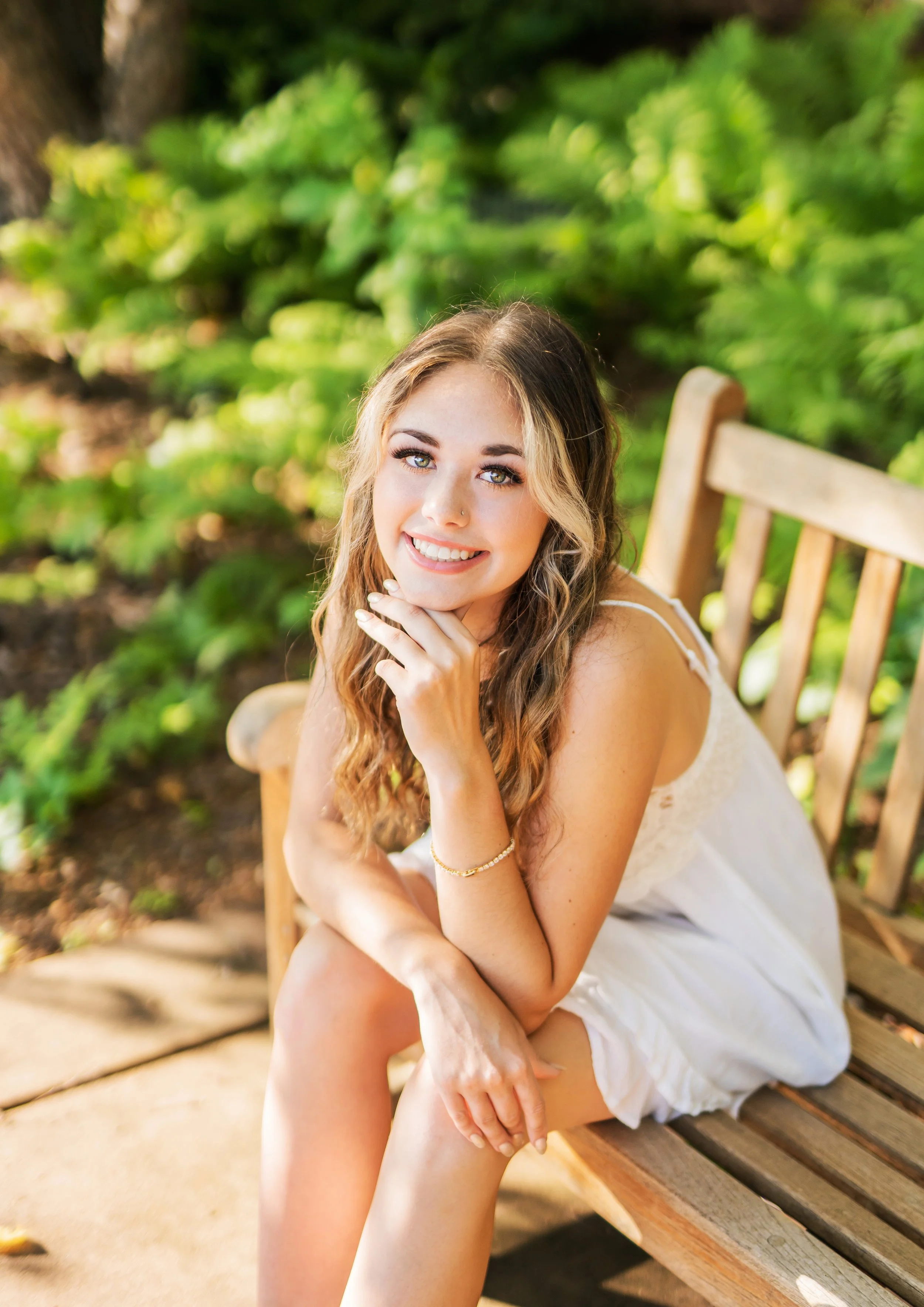A young woman with long, wavy hair and a white dress sitting on a wooden park bench, smiling and looking at the camera, surrounded by greenery.