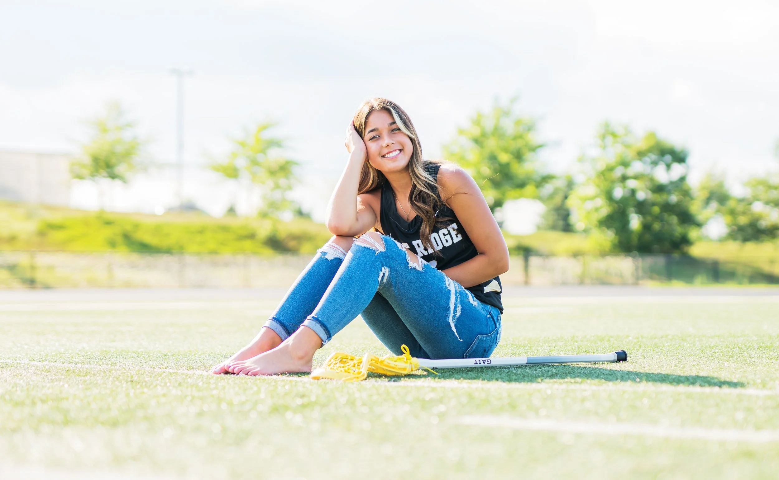 Young woman sitting on a sports field, smiling, with sports equipment nearby, wearing ripped jeans and a sleeveless shirt.