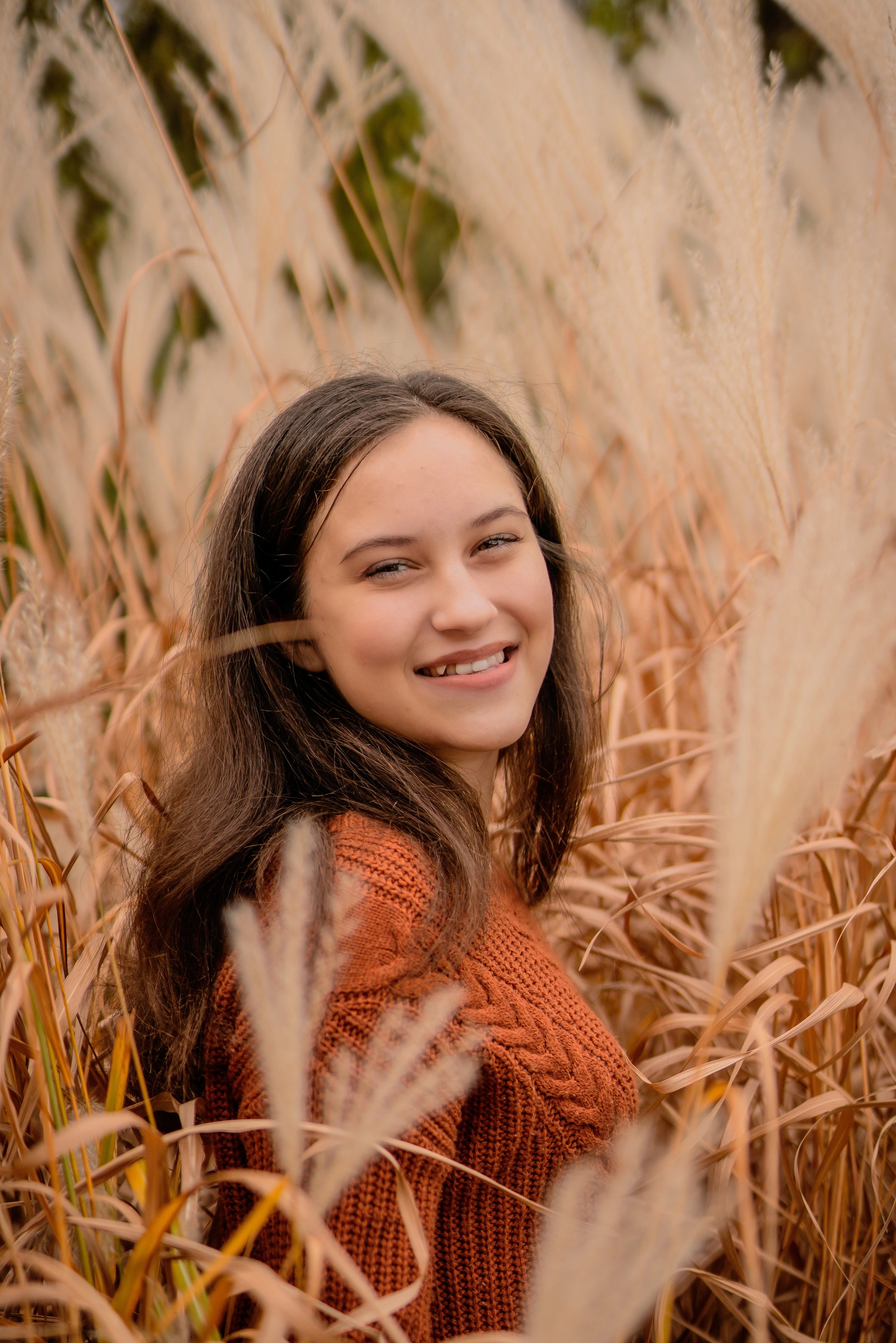 A young woman with brown hair smiling and wearing an orange sweater standing among tall golden grass.