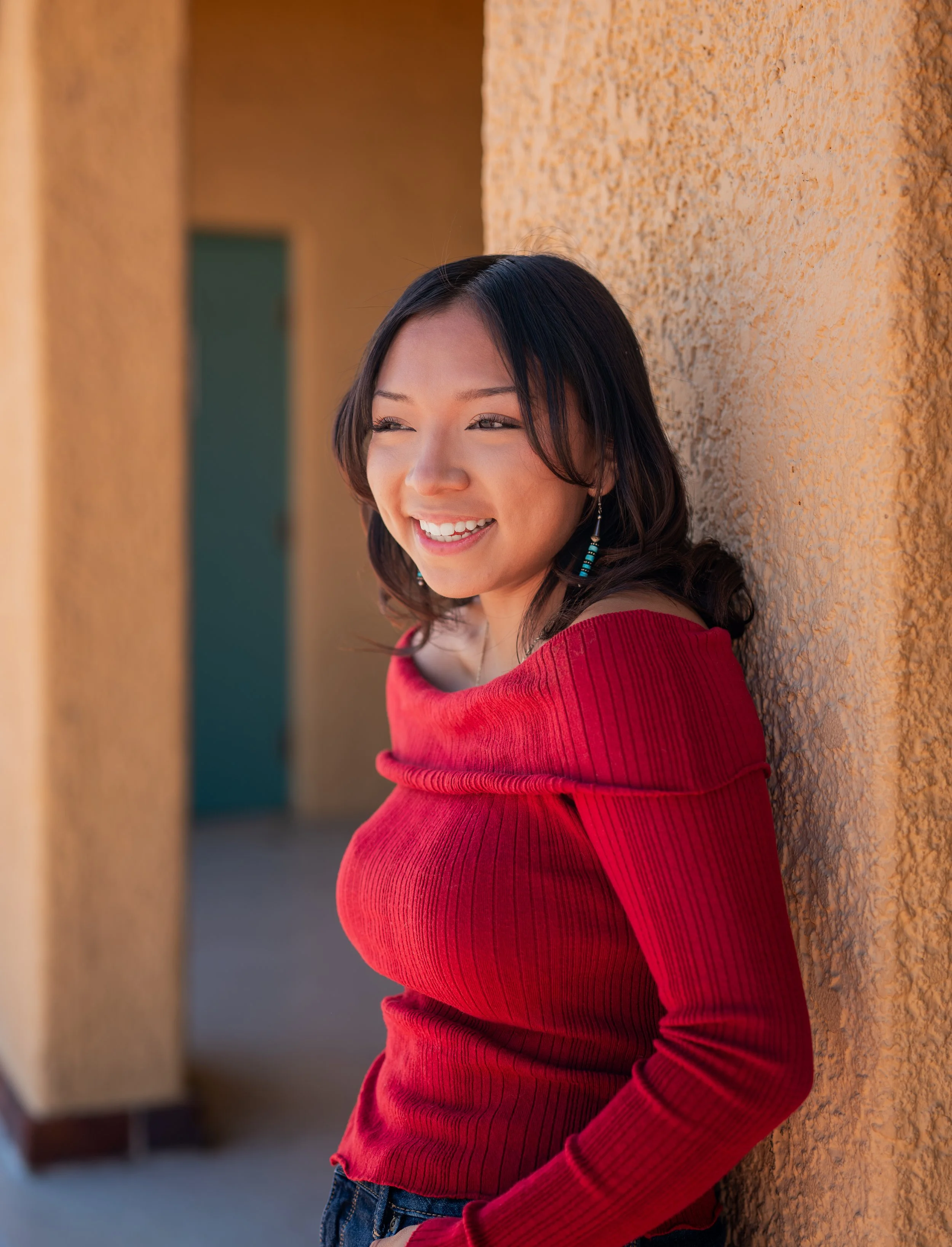 A woman with dark hair smiling while leaning against a textured beige wall, wearing a red long-sleeve top and earrings, with a blurred background.