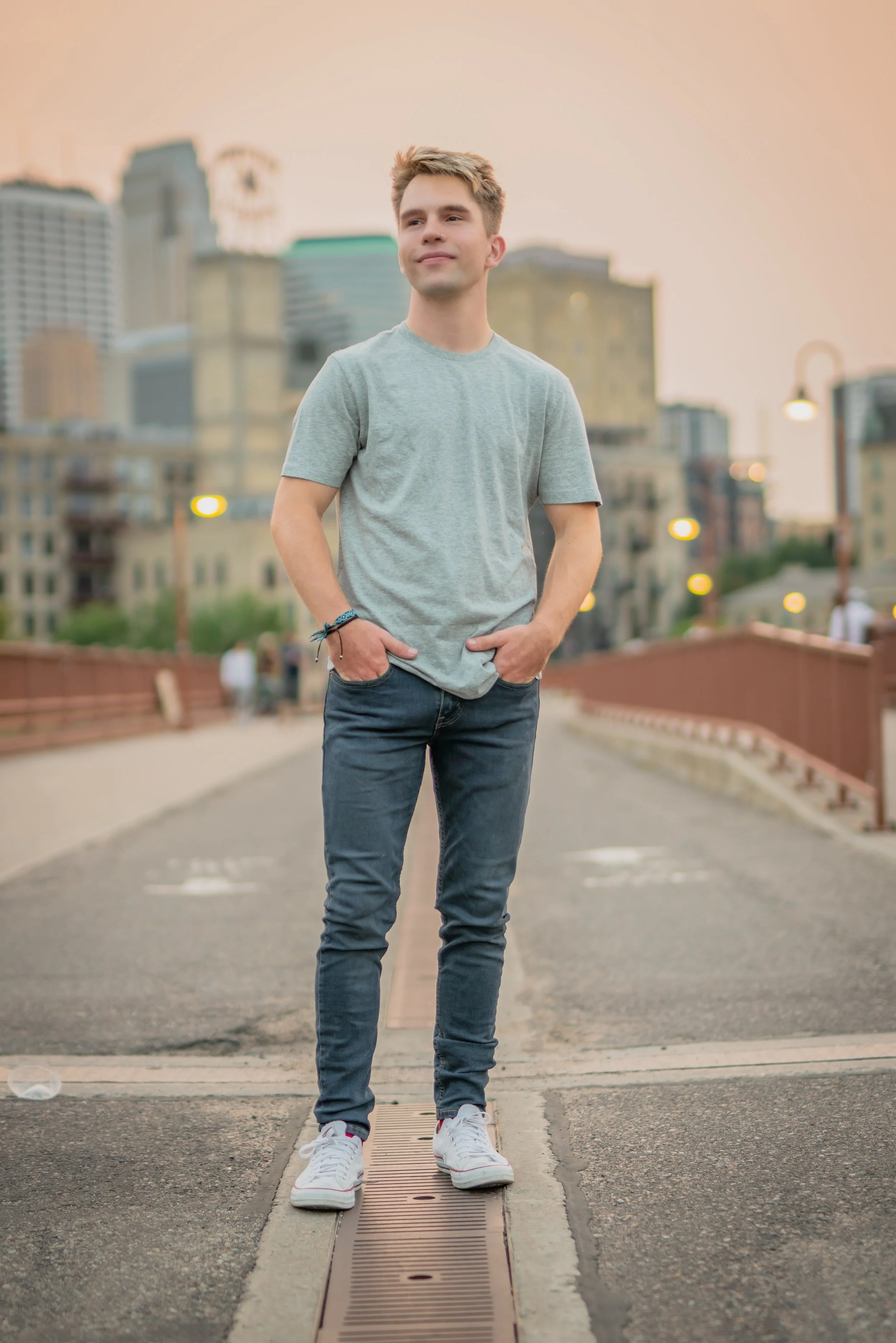 A young man standing on a pedestrian bridge in an urban area at sunset, wearing a gray t-shirt, jeans, and white sneakers, with a city skyline in the background.
