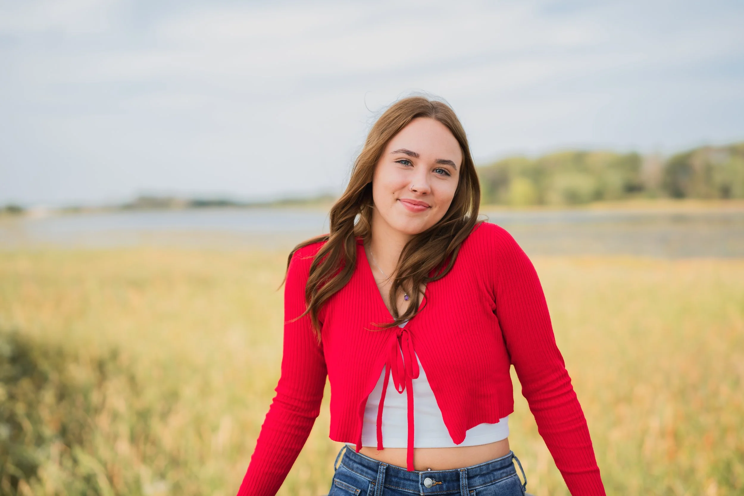 A young woman with long brown hair standing outdoors in a field with a body of water and trees in the background, wearing a red cropped cardigan and jeans, smiling at the camera.