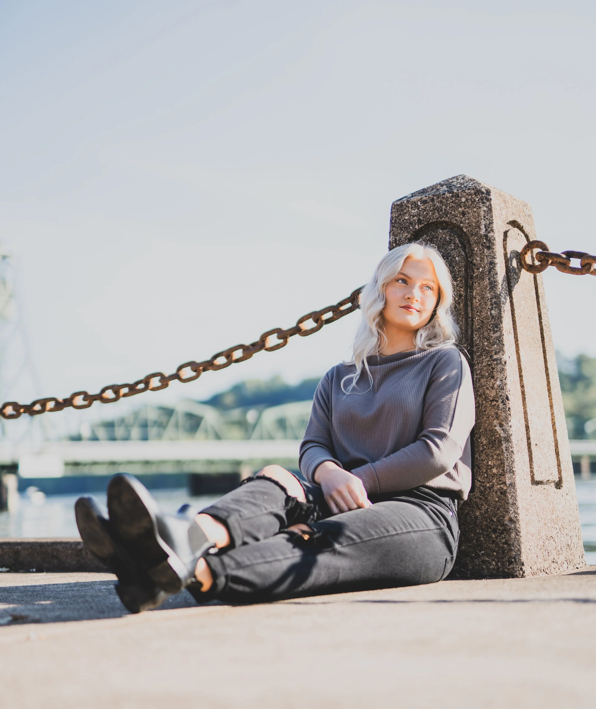 A young woman with platinum blonde hair sitting on a concrete pier near water, leaning against a concrete post with a chain, looking thoughtfully into the distance on a clear day.