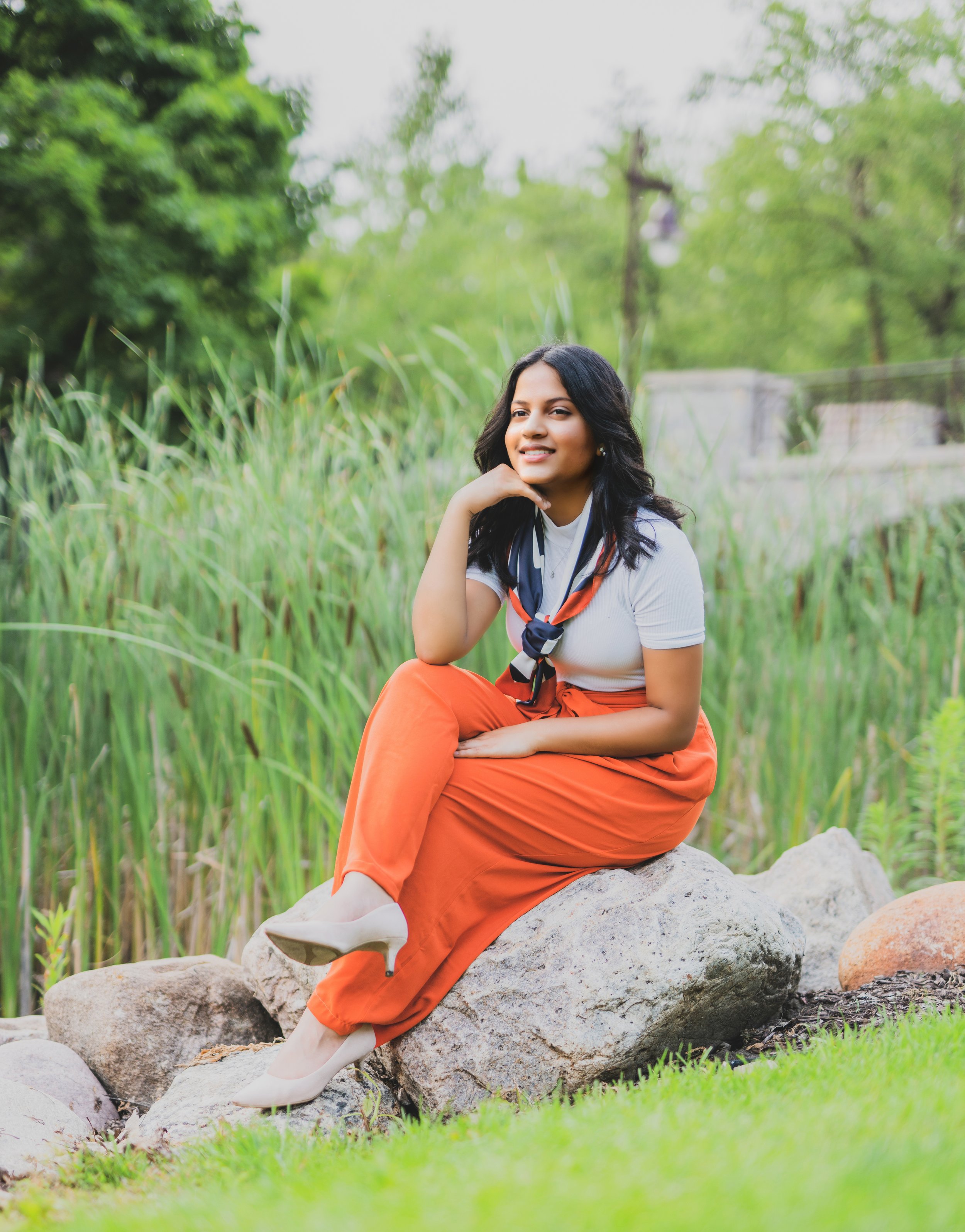 A woman sitting on a large rock in a green outdoor park setting, wearing orange pants, a white top, and a navy scarf, with a background of tall grasses, trees, and a few lampposts.