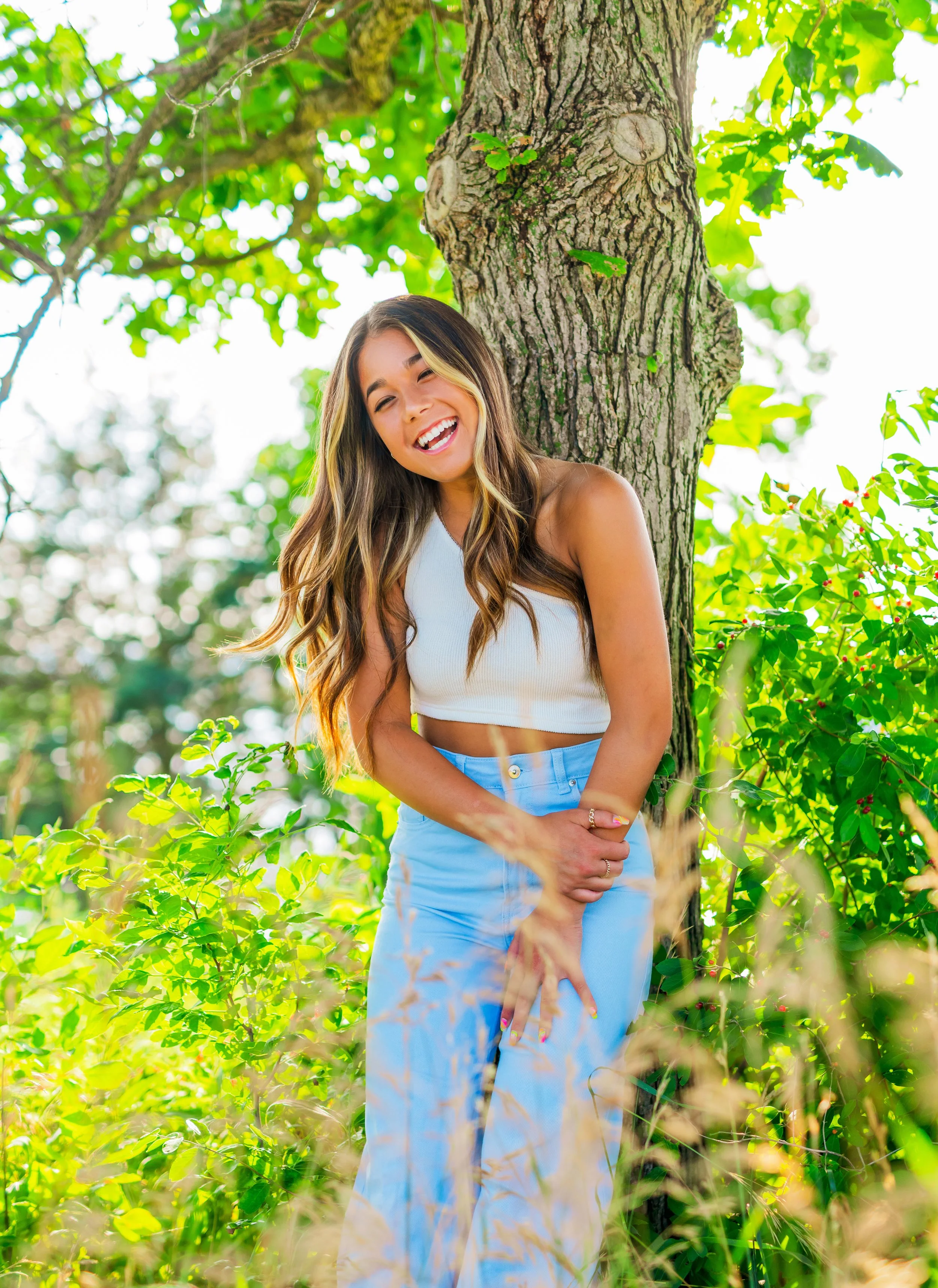 Young woman with long wavy hair smiling and laughing while standing outdoors next to a tree amid green foliage, wearing a white sleeveless top and light blue pants.