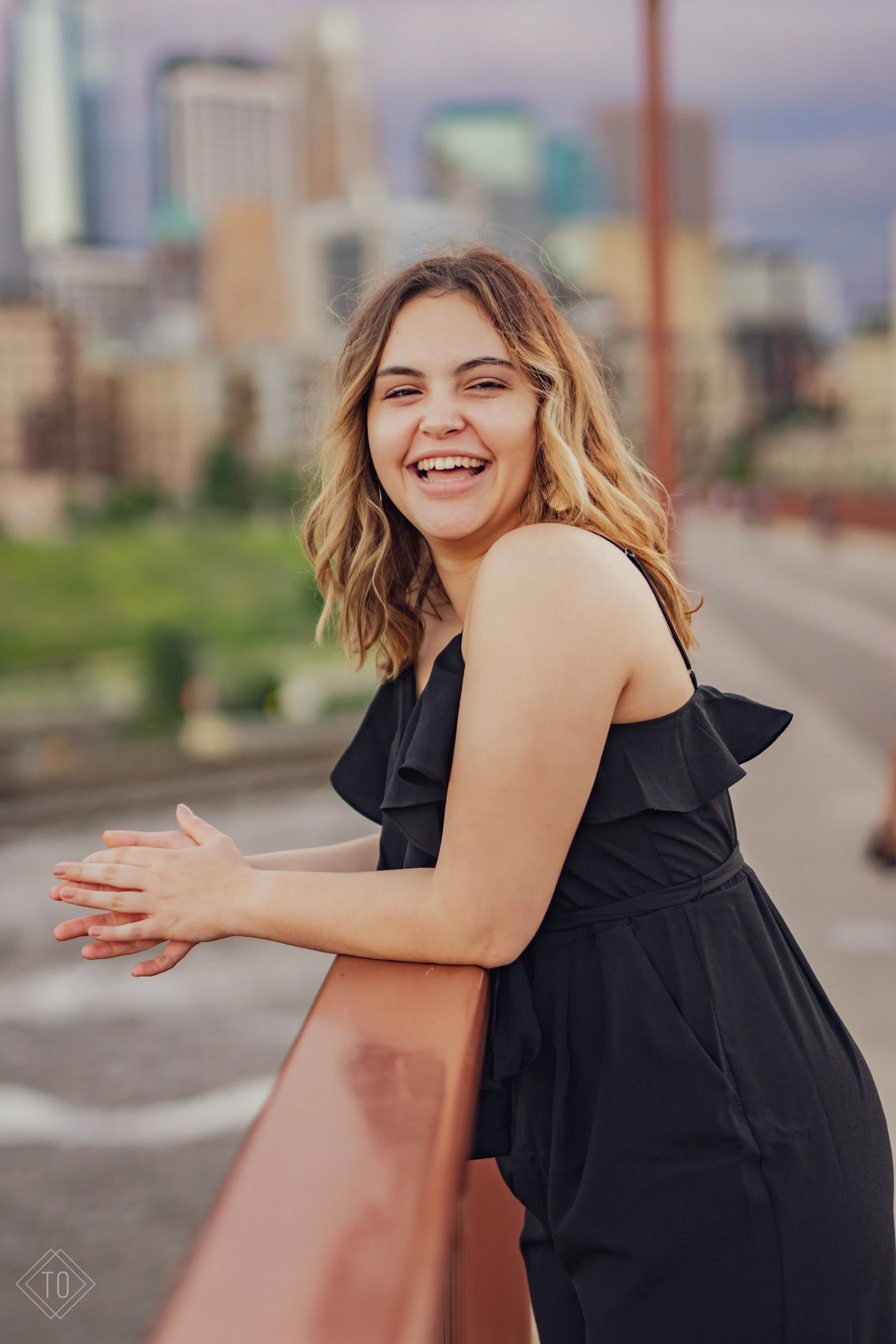 A young woman with shoulder-length wavy hair smiling and laughing while leaning on a bridge railing with a cityscape in the background.