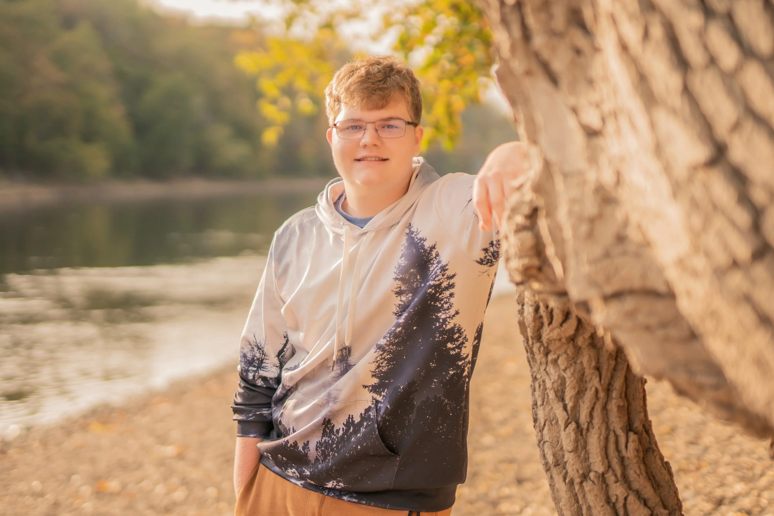 A young man with glasses standing outdoors beside a tree near a river, with autumn foliage in the background.