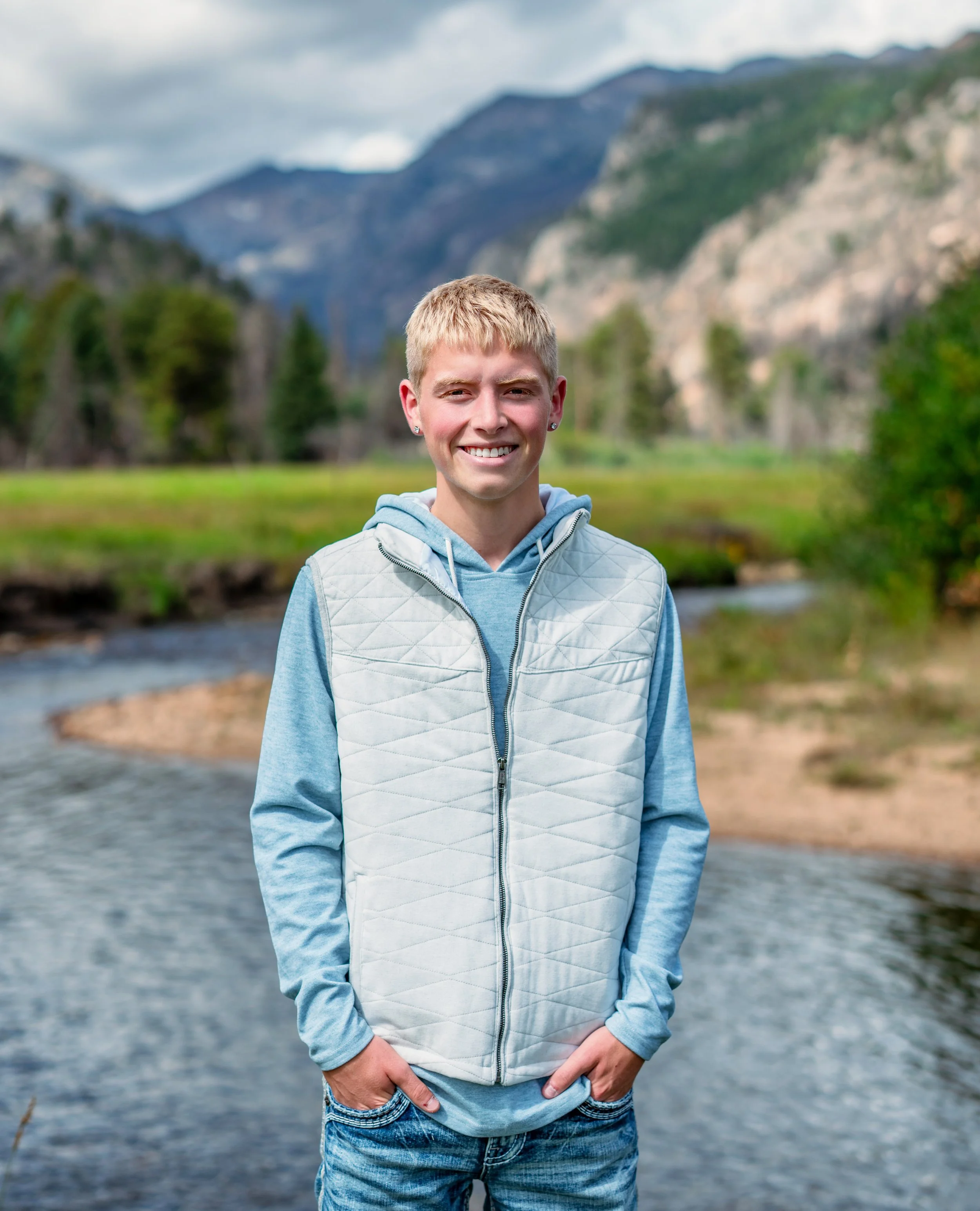 A smiling young man with blonde hair standing outdoors near a river, with mountains, trees, and cloudy sky in the background.