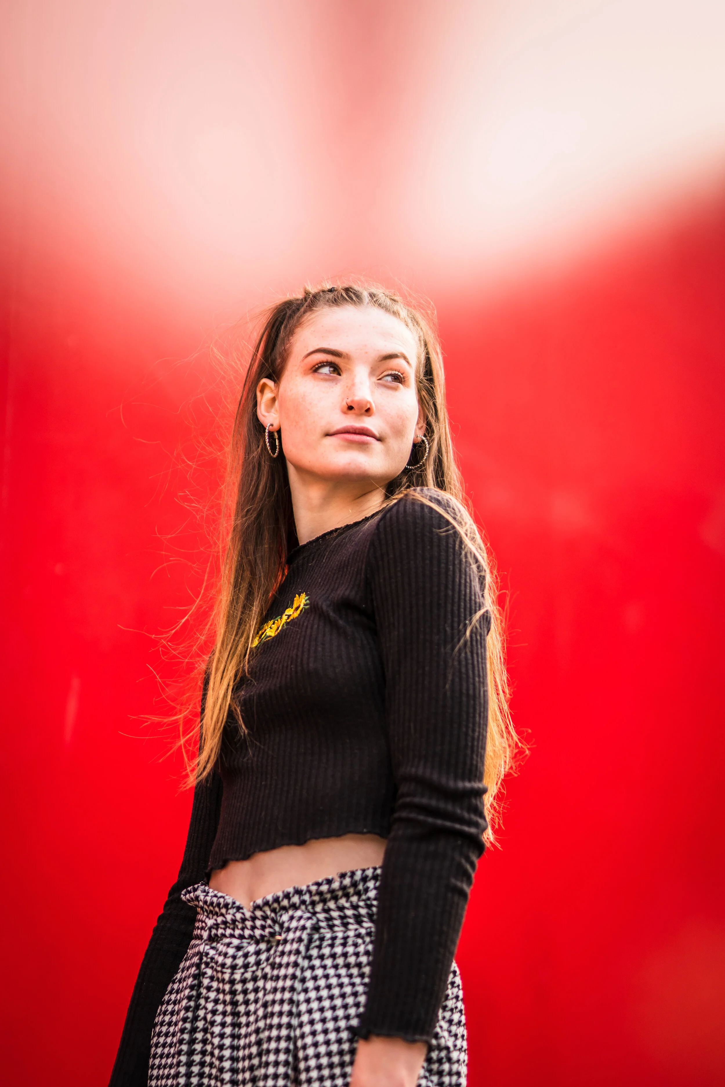 A young woman with long brown hair standing outdoors against a bright red background, wearing earrings, a black cropped long-sleeve top, and black and white checkered pants.