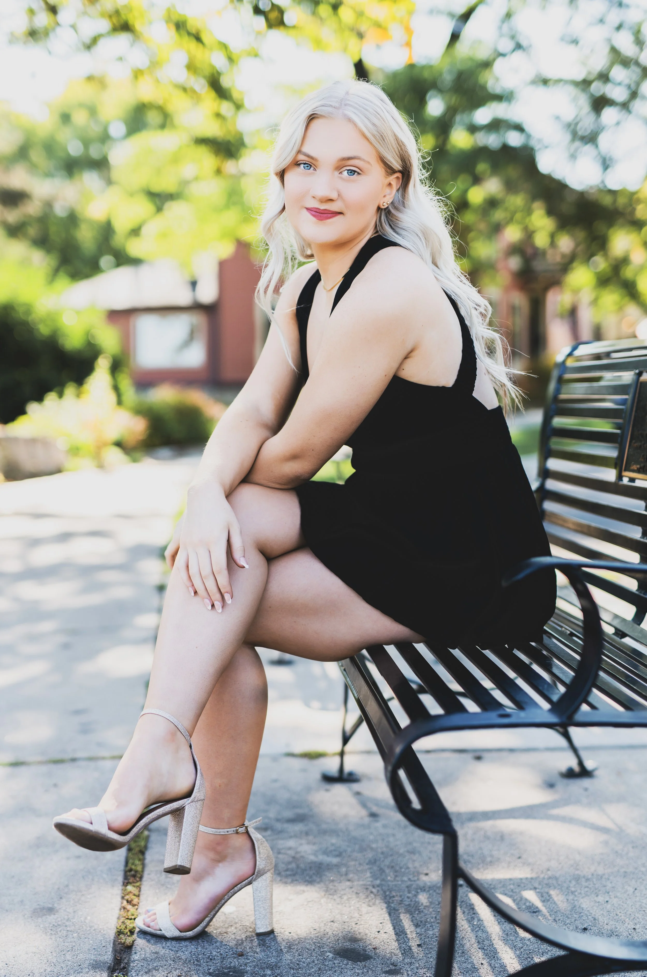 Young woman with blonde hair and blue eyes, smiling, sitting on a bench outdoors. She is wearing a black dress and beige high-heeled sandals, with her legs crossed.