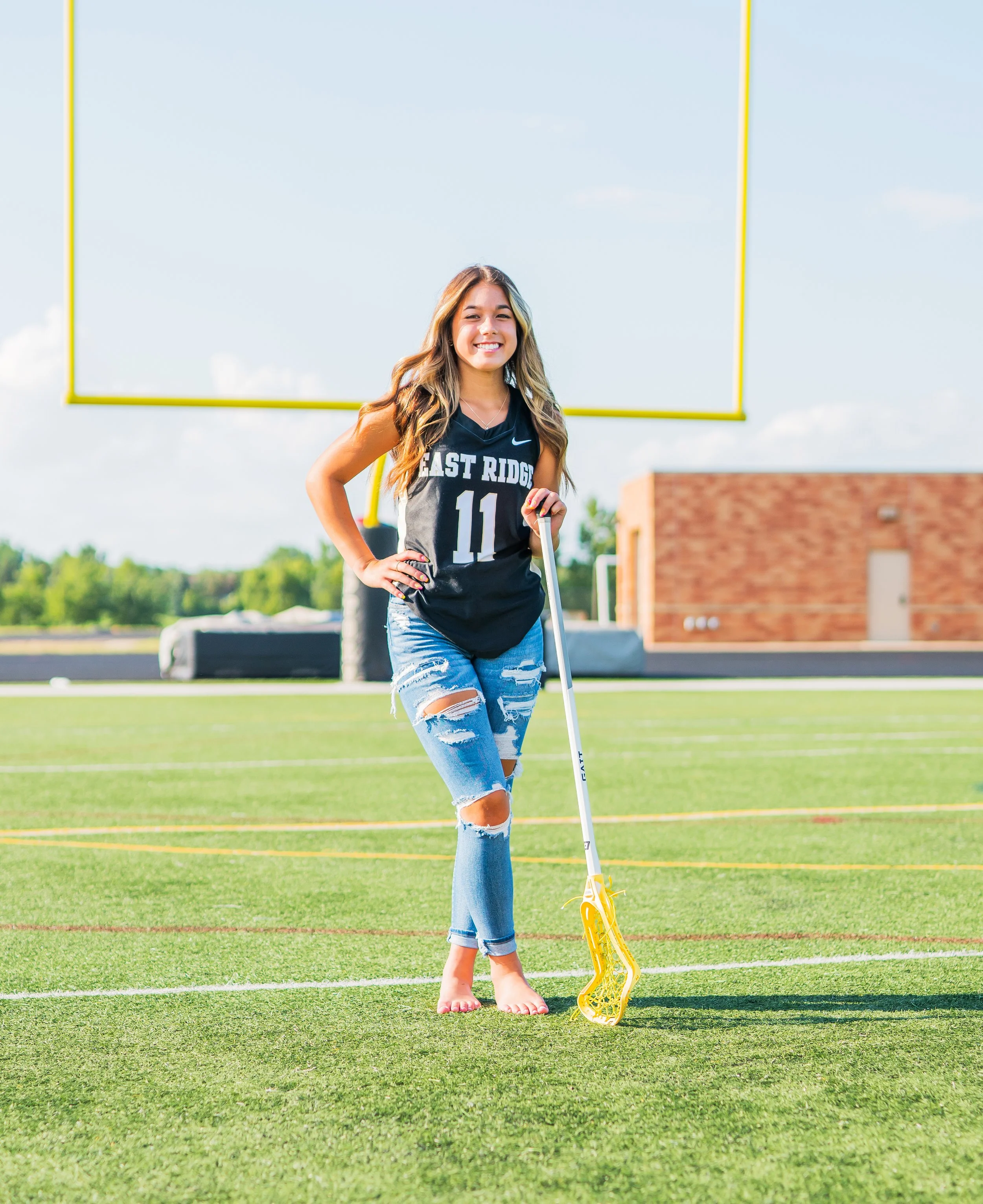 Young woman smiling on a football field holding a lacrosse stick, wearing a sports jersey and ripped jeans.