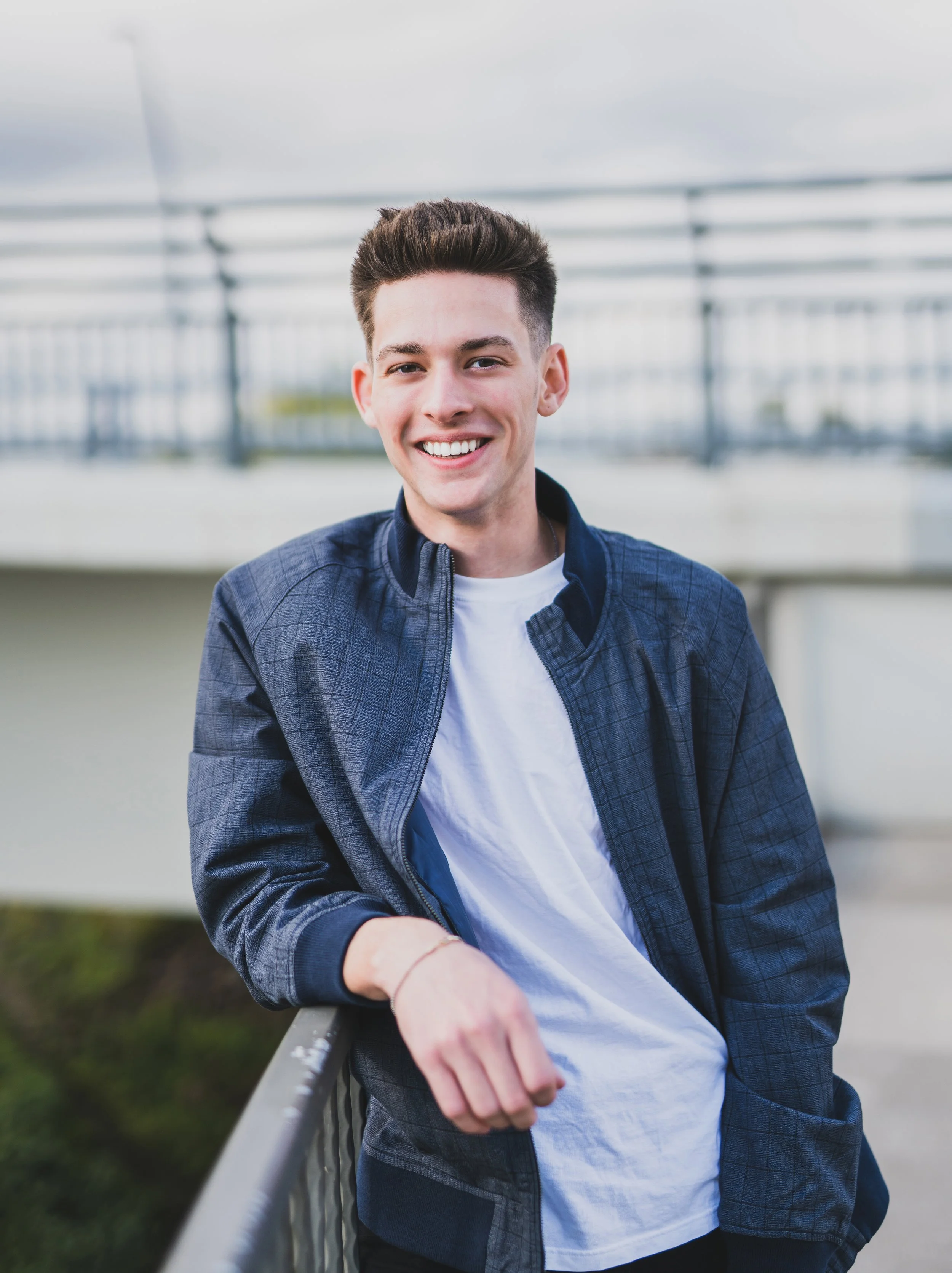Young man smiling outdoors, wearing a navy blue jacket and white T-shirt, standing by a metal railing with a blurred background of an outdoor setting.