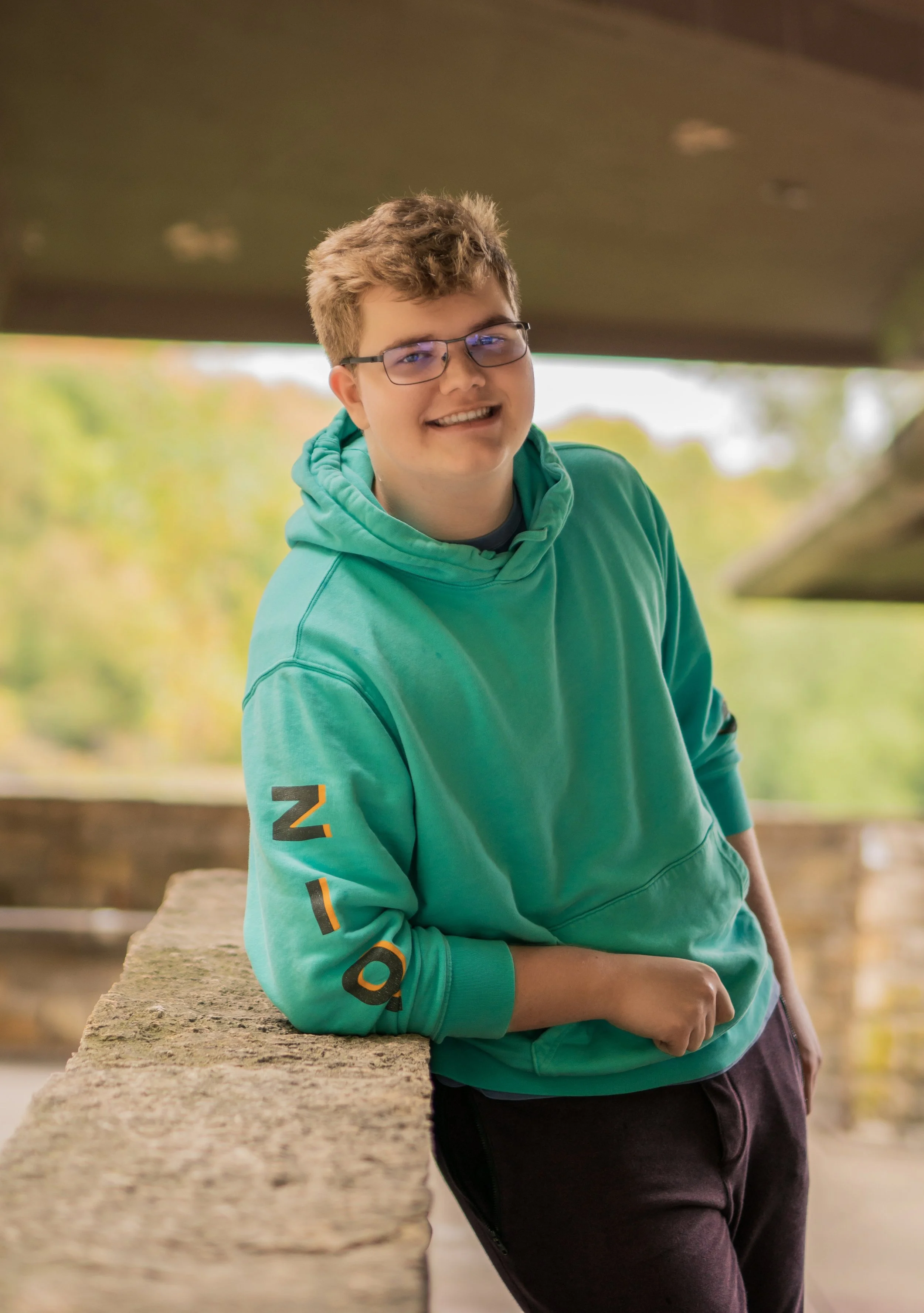 A young man with glasses and short, curly hair leaning on a stone railing outdoors, smiling at the camera, wearing a turquoise hoodie with the word 'ICON' on the sleeve, and brown pants, with a background of blurred green trees.
