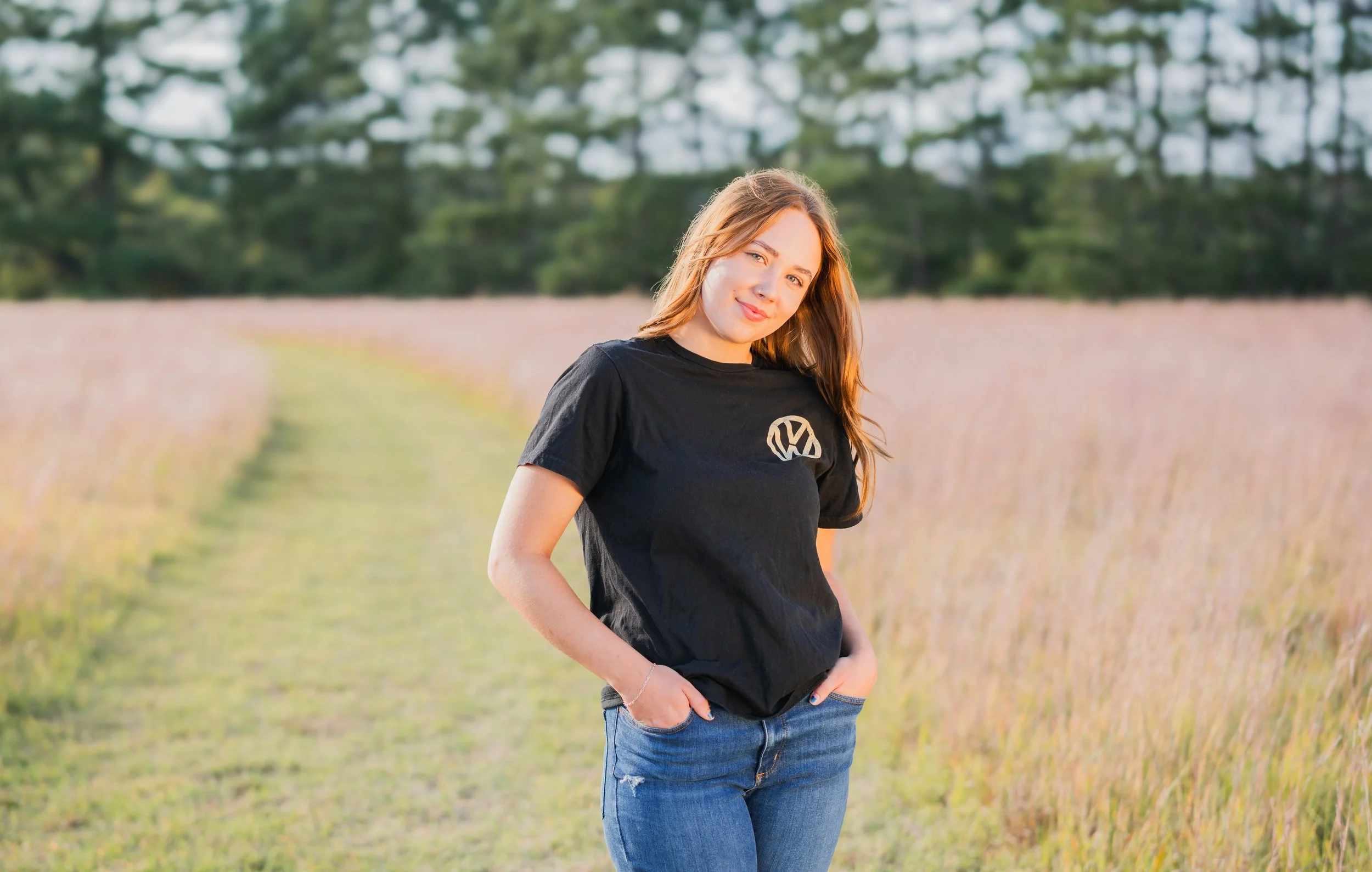 A young woman with long red hair stands in a field during golden hour, wearing a black T-shirt with a Volkswagen logo and blue jeans, smiling slightly with her hands in her pockets, with trees in the background.
