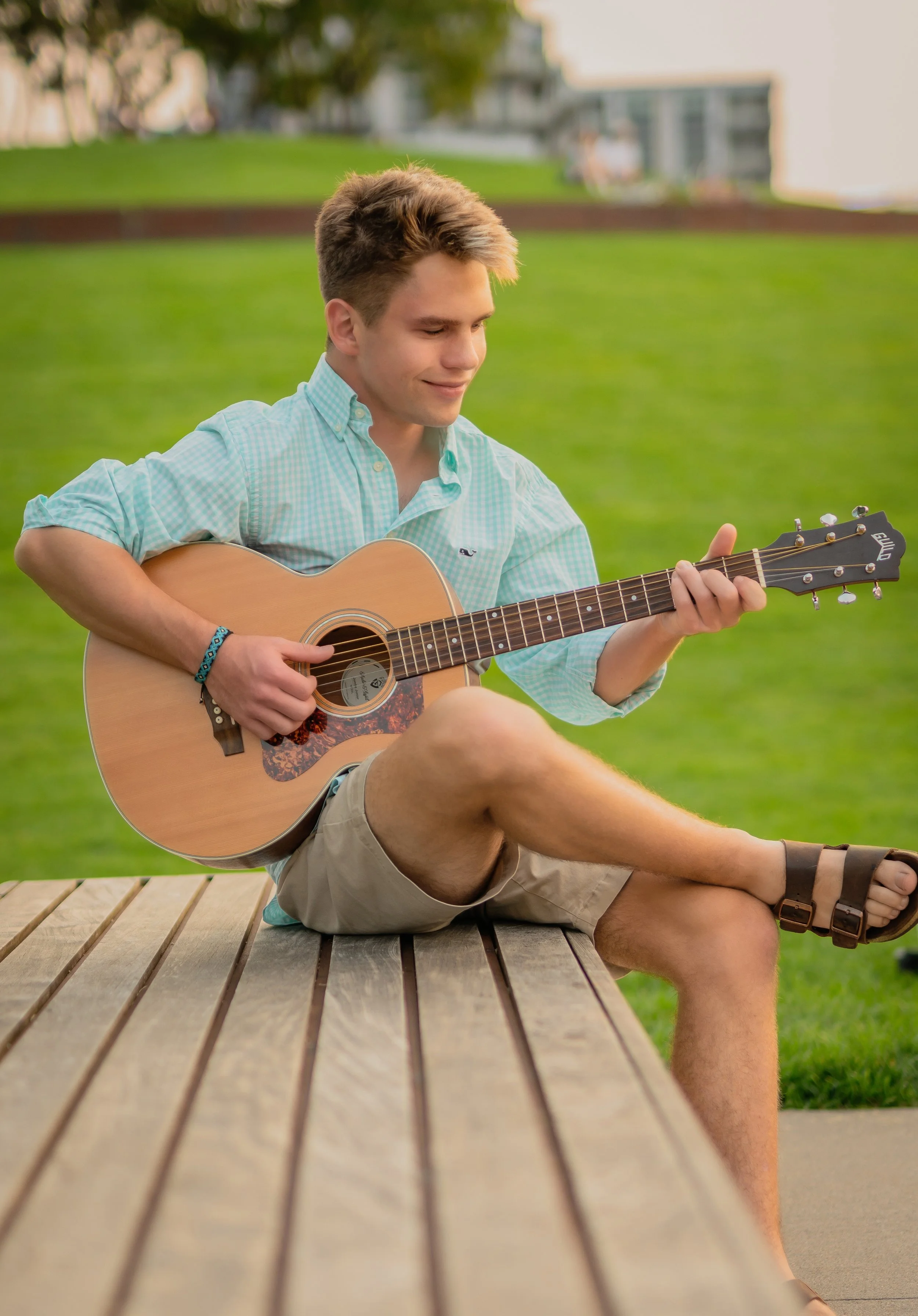 A young man sitting on a wooden bench outdoors, playing an acoustic guitar with a green grassy background.