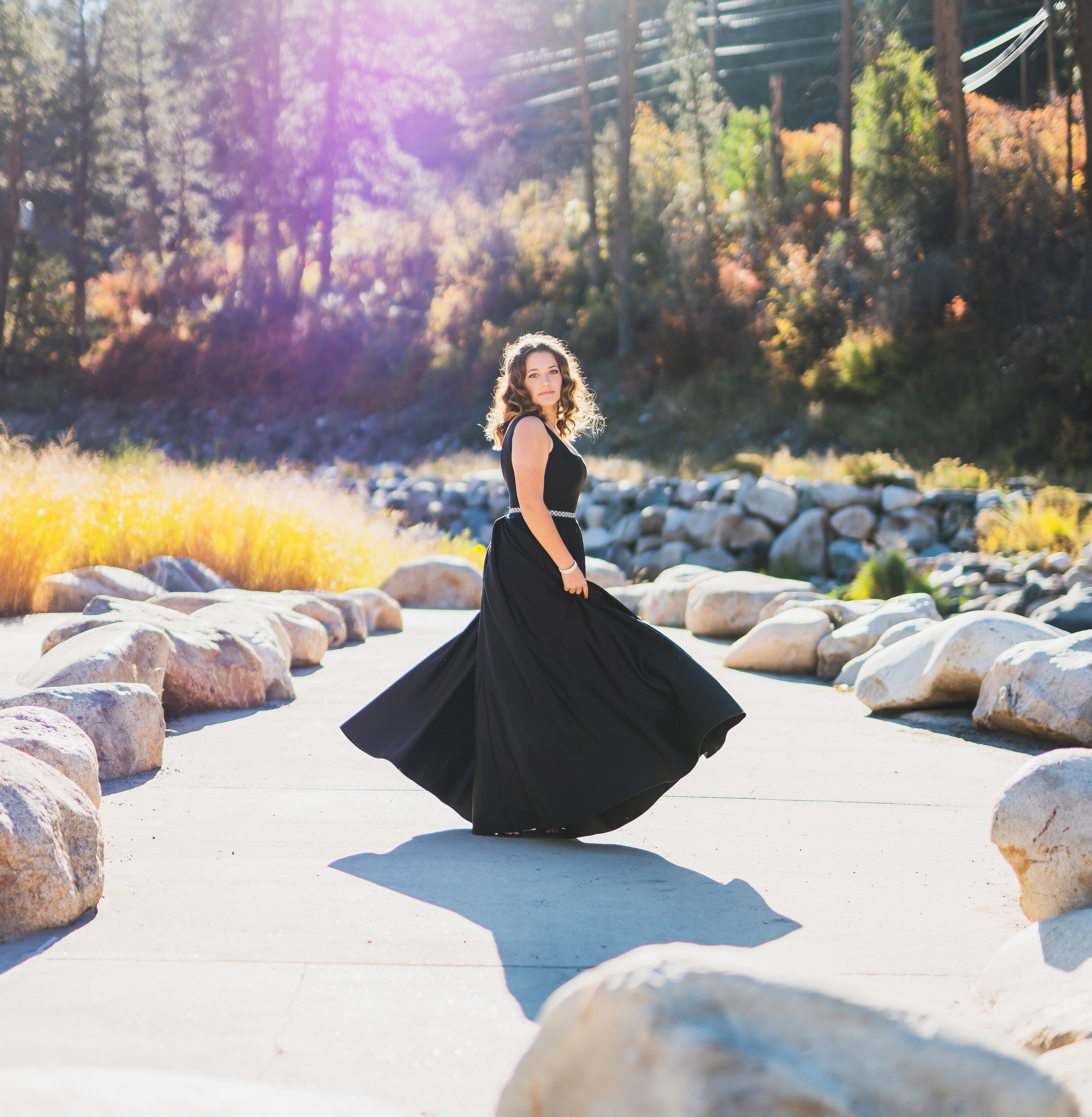 A woman in a long black dress twirling outdoors on a sunny day, surrounded by rocks and trees with autumn foliage.