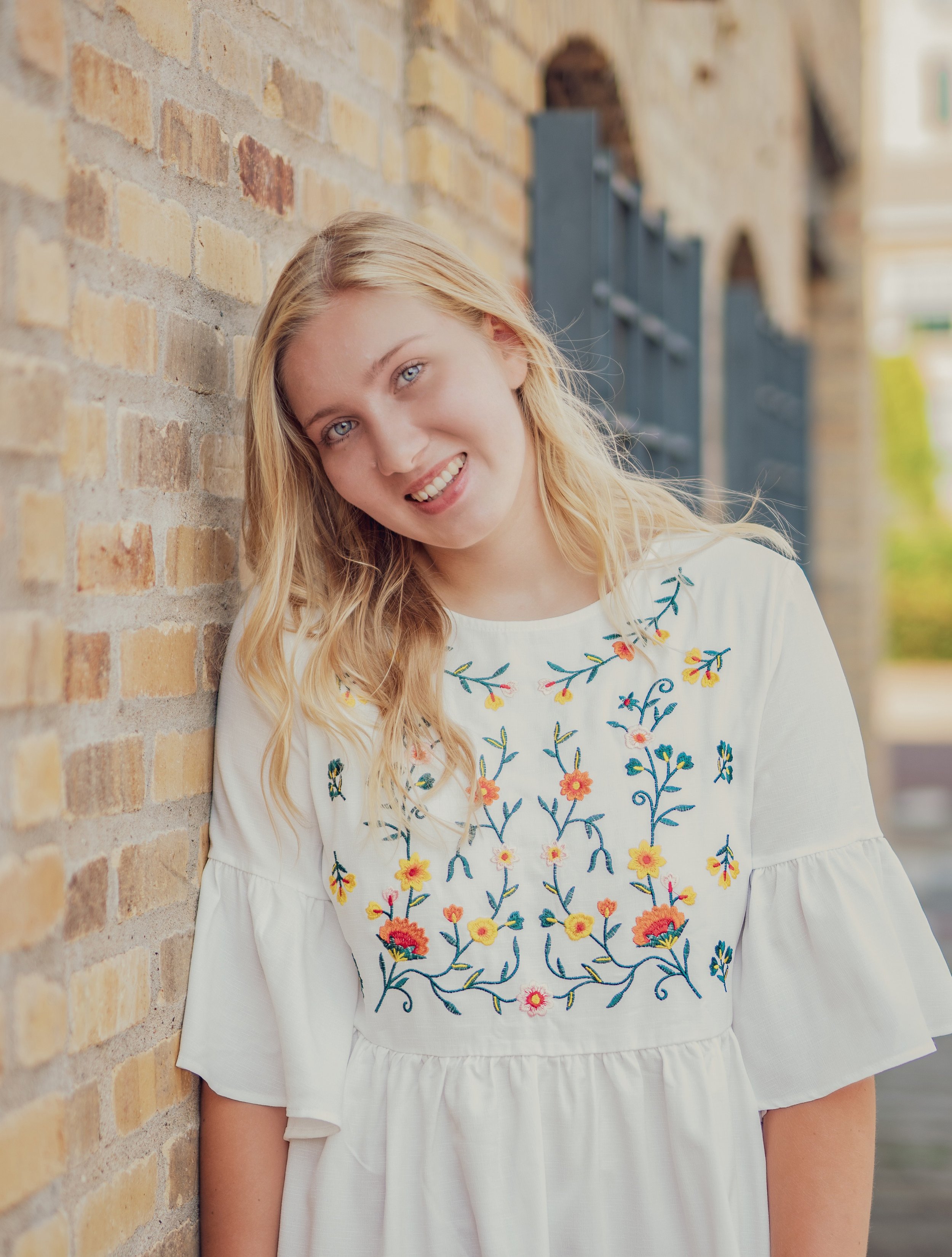 A young woman with blond hair and blue eyes smiling while leaning against a brick wall, wearing a white embroidered dress with floral patterns.