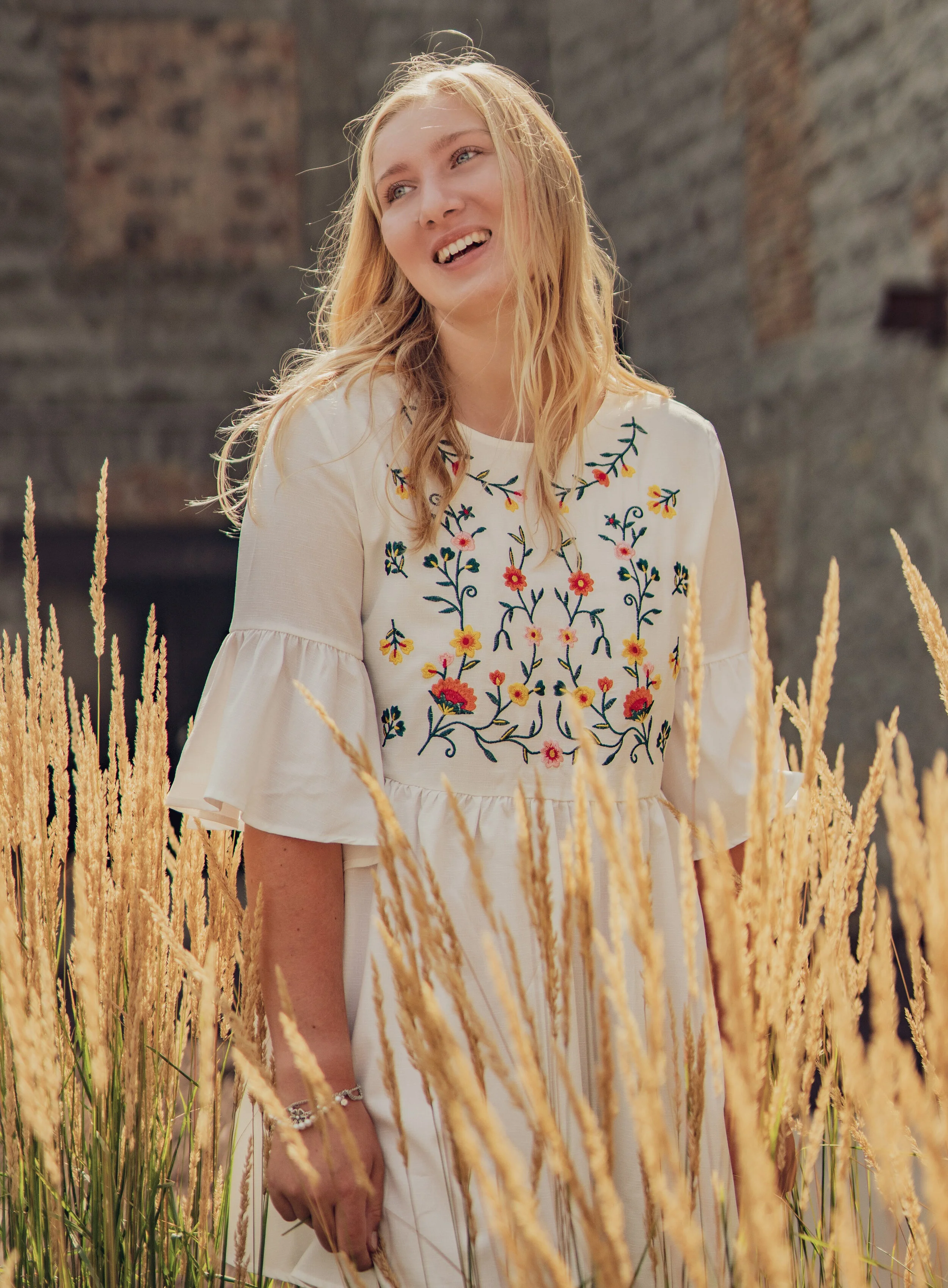 A young woman with blonde hair wearing a white embroidered dress stands among tall, golden grasses in an outdoor setting with a brick building in the background.