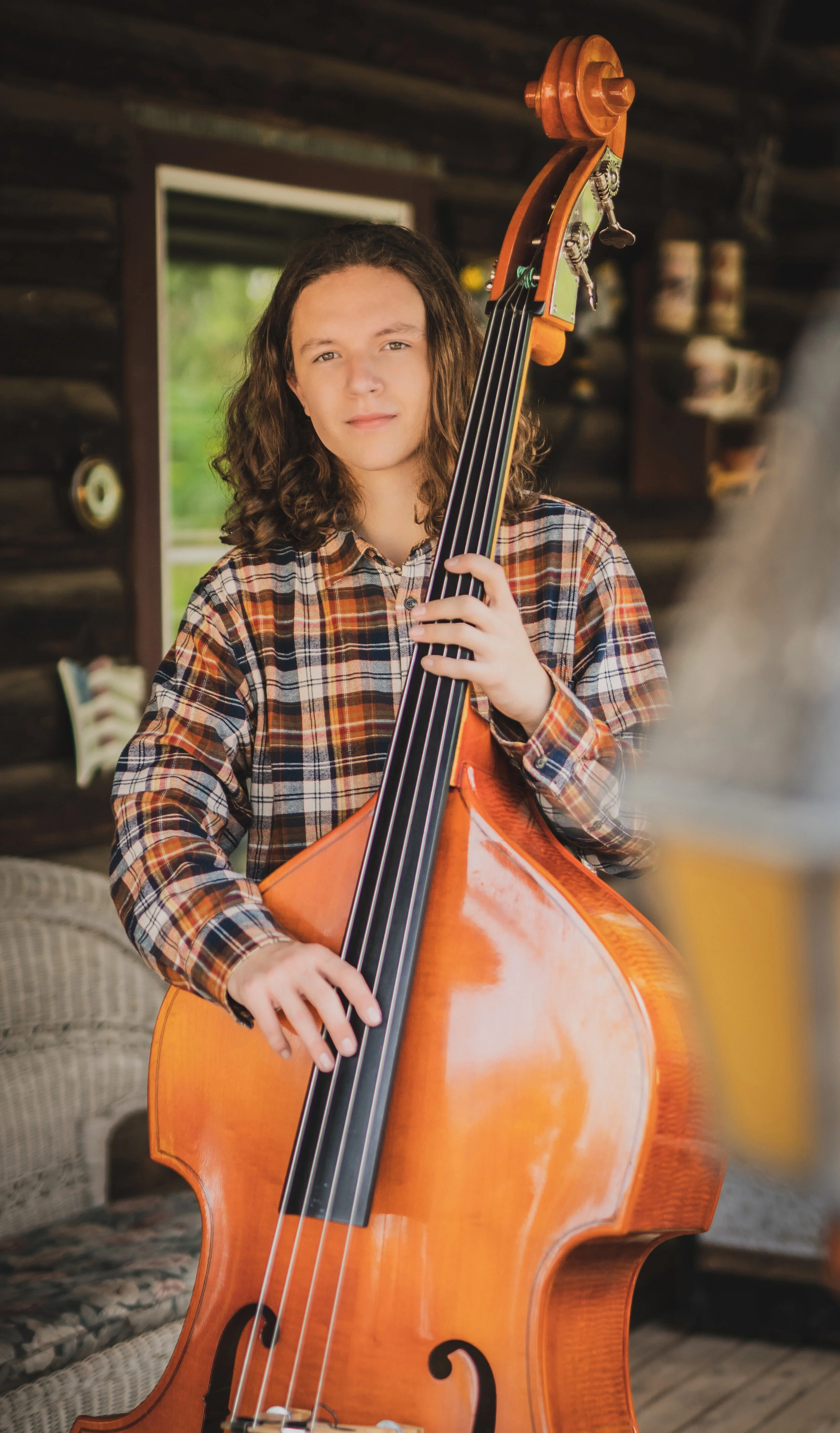 A young person with shoulder-length curly hair in a plaid shirt standing in a rustic room, holding a large upright bass guitar.