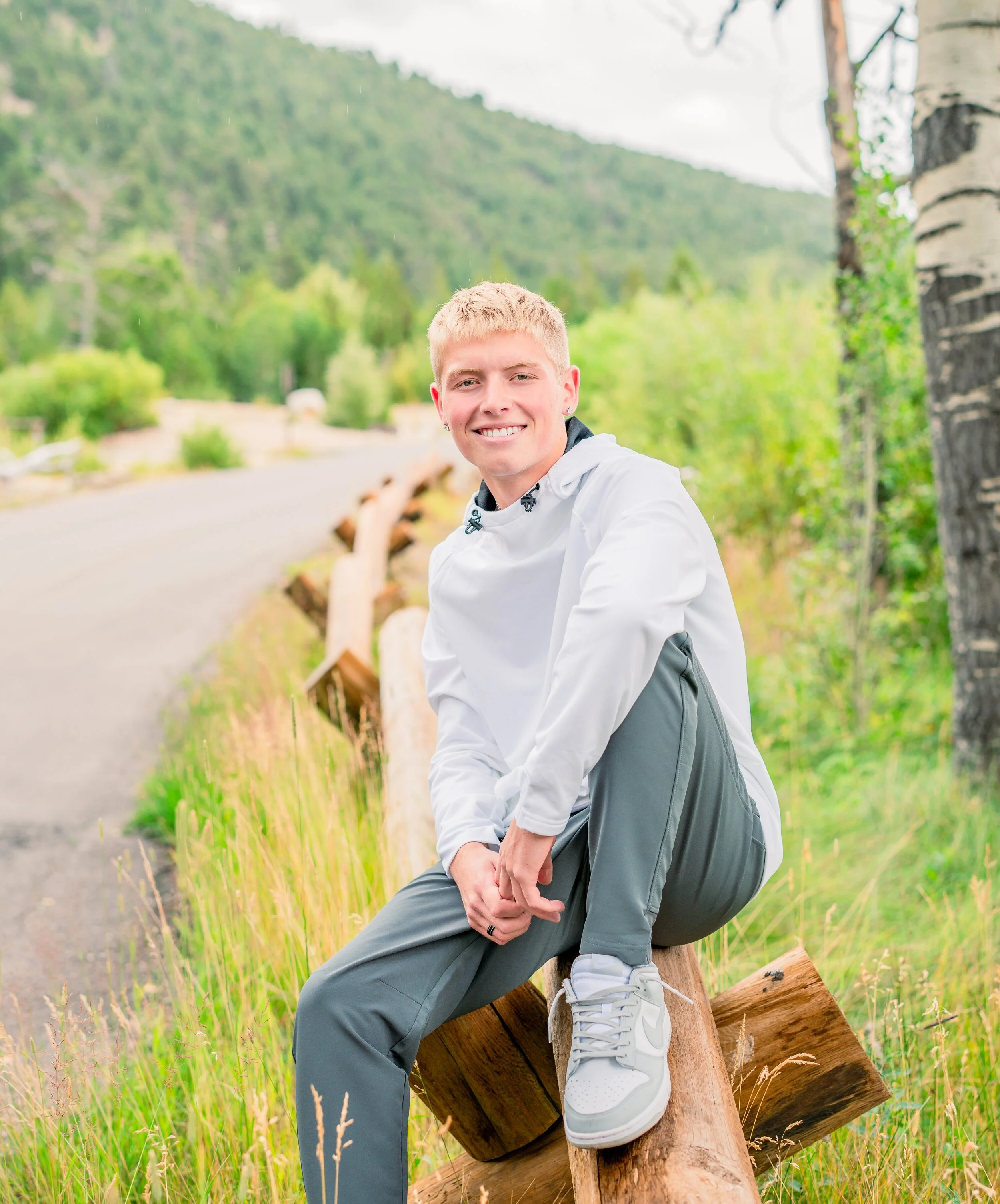 A young man with blonde hair and a white jacket sitting on a fallen log beside a road in a scenic green mountain area, smiling at the camera.