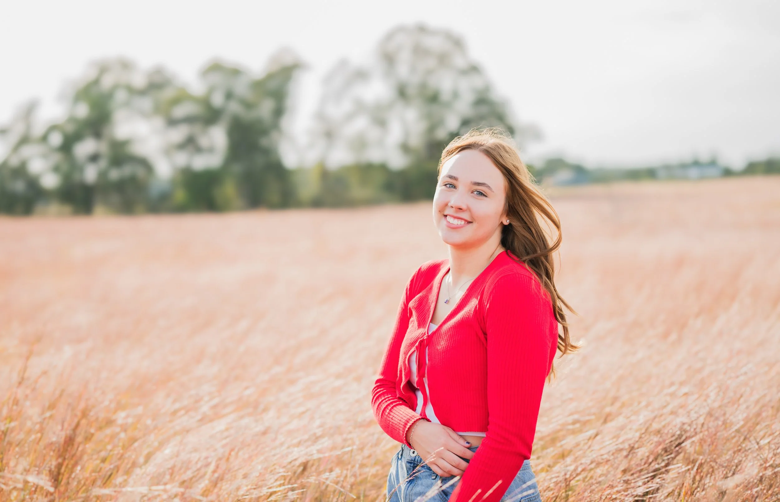 A young woman with long, wavy hair standing in a field of tall, golden grass, smiling, wearing a red long-sleeve top and earrings.