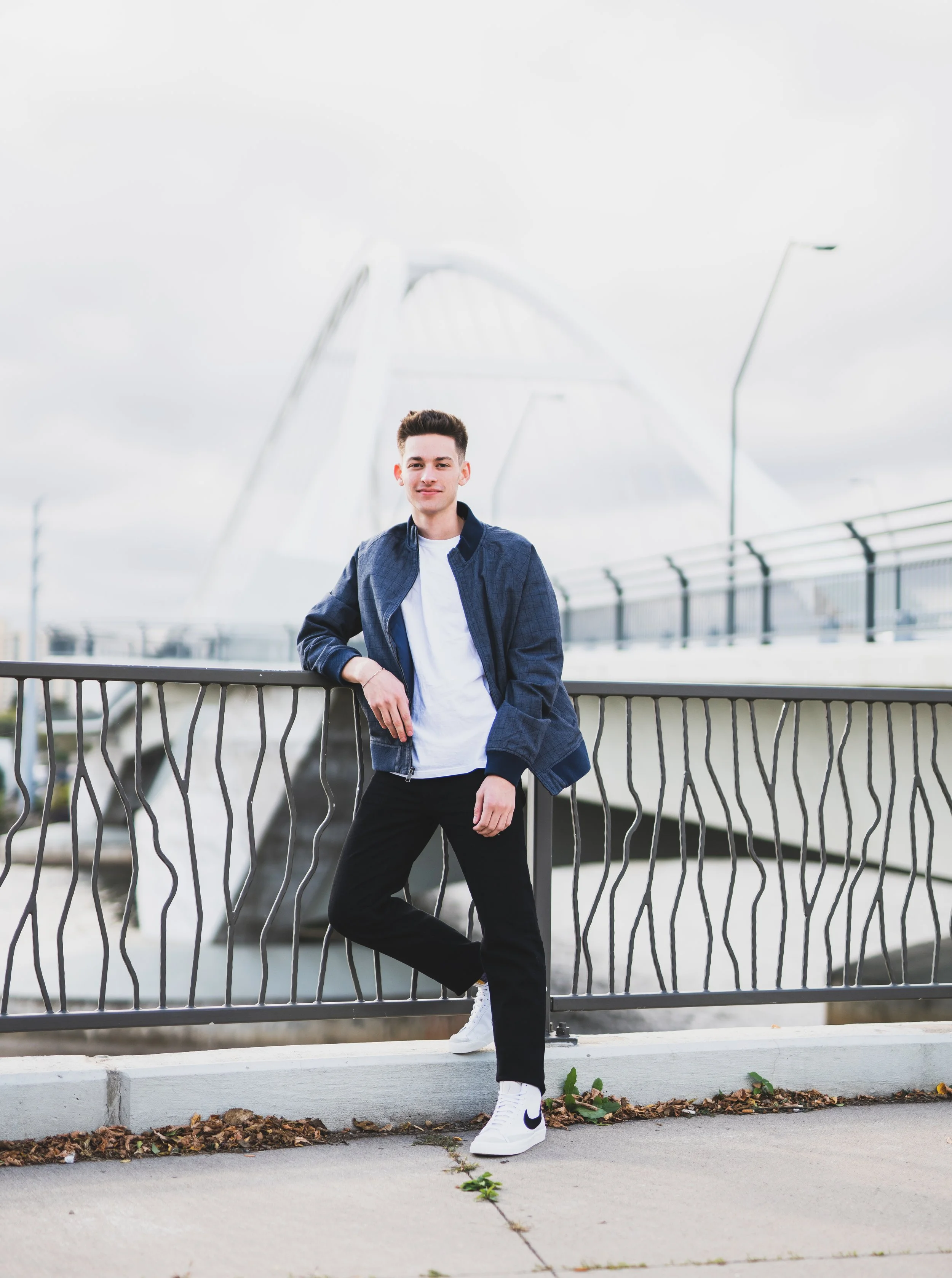 A young man with short dark hair, wearing a denim jacket, white T-shirt, black pants, and white Nike sneakers, standing outdoors on a sidewalk near a metal railing with a modern bridge structure in the background under a cloudy sky.