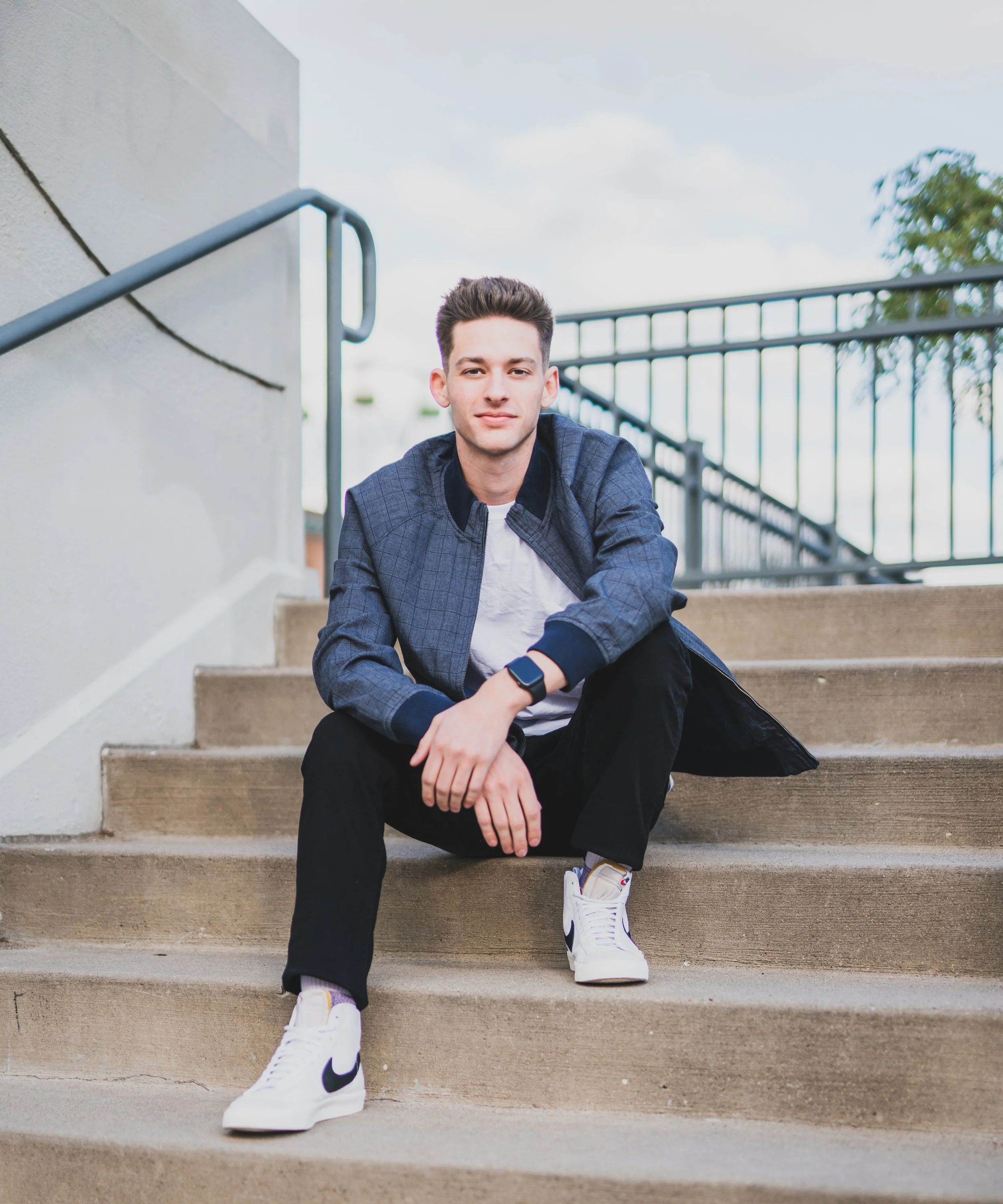 Young man sitting on outdoor stairs with a railing, wearing a dark blue jacket, white t-shirt, black pants, and white Nike sneakers, looking at the camera with a slight smile, cloudy sky in the background.