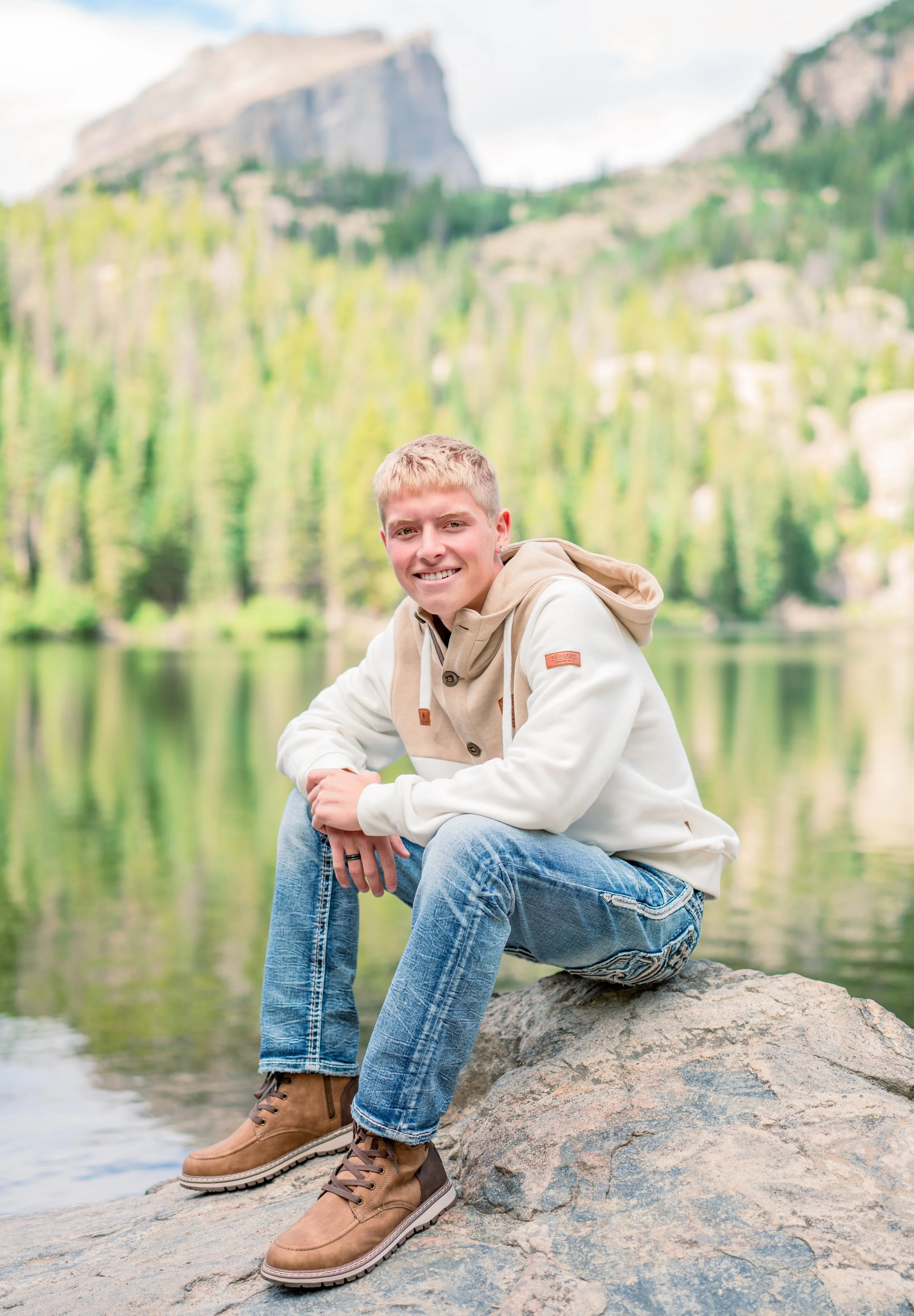 A young man with blond hair, wearing a beige and white hoodie, blue jeans, and tan boots, sitting on a large rock by a lake surrounded by green trees and mountains in the background, smiling at the camera.