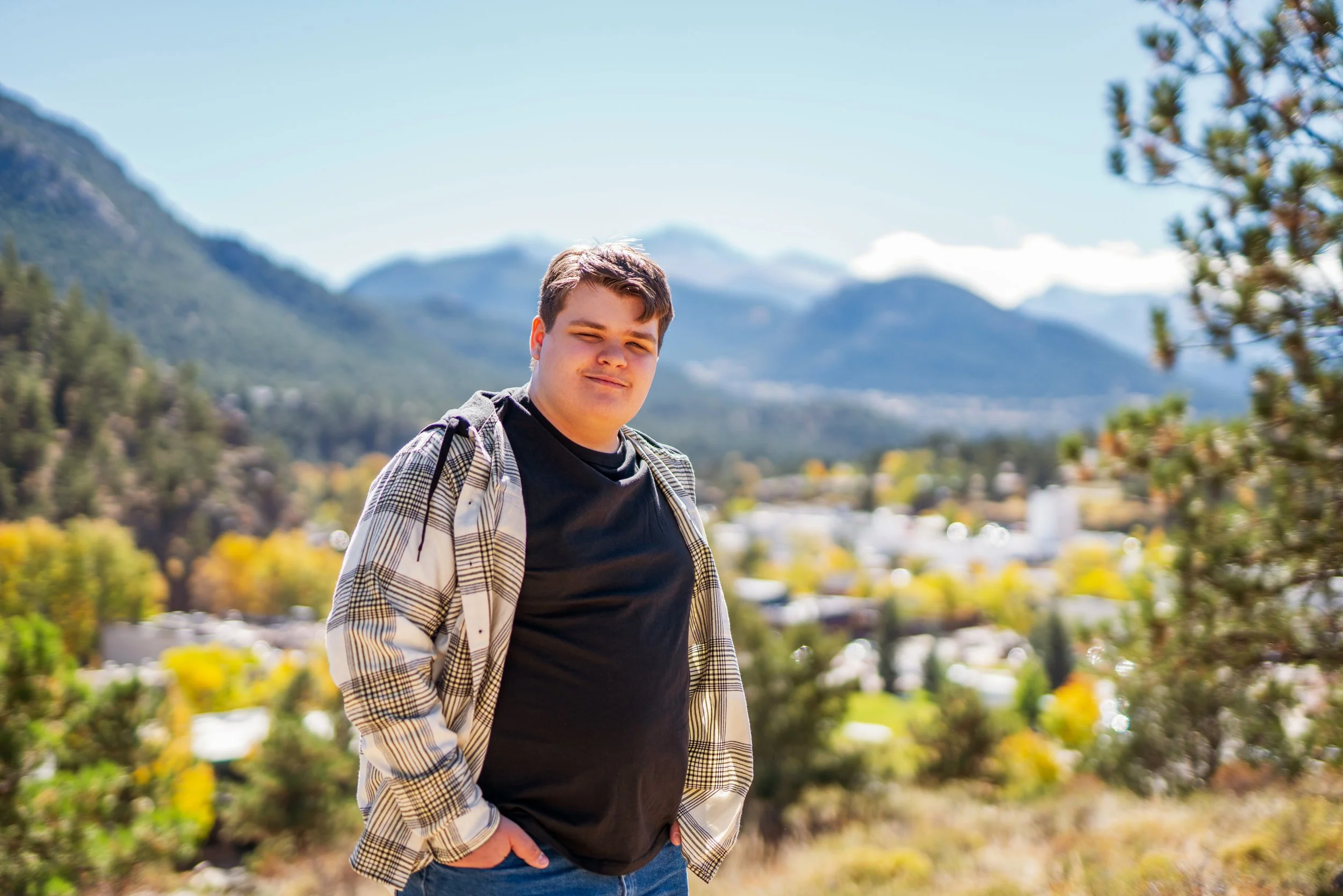 A young man outdoors with mountains and trees in the background, wearing a black T-shirt and a plaid shirt, with his hands in his pockets, smiling slightly.