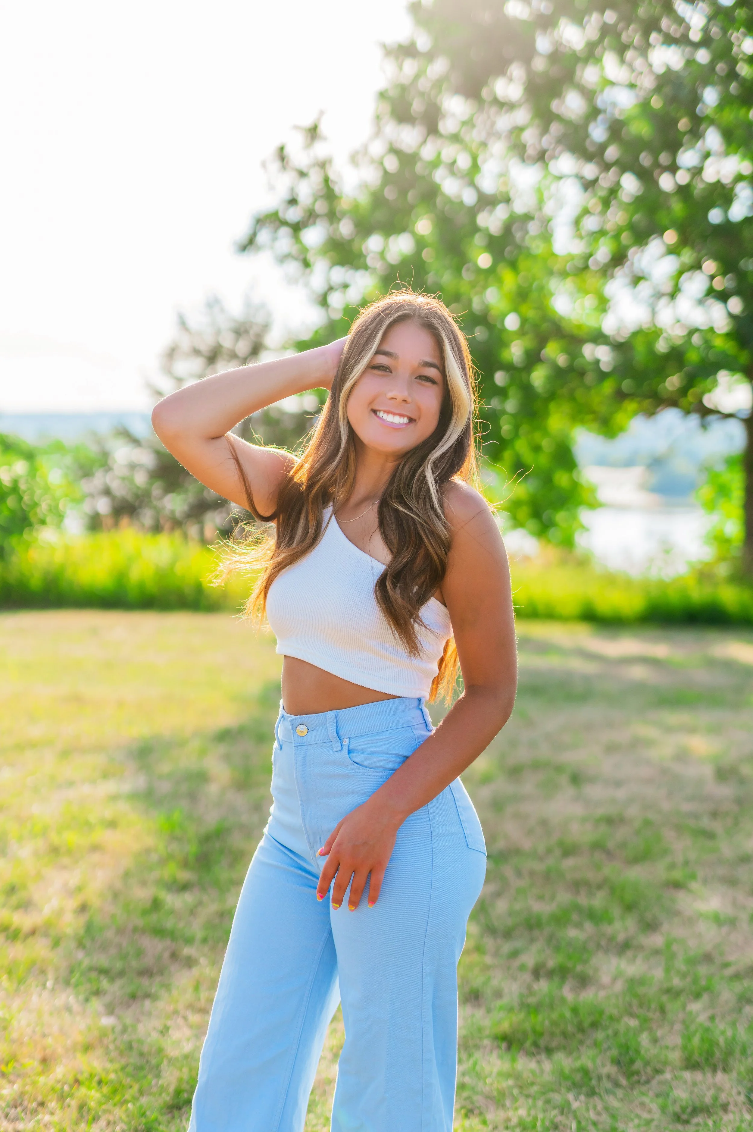 A young woman with long wavy hair smiling outdoors in a sunny park, wearing a white crop top and light blue jeans.
