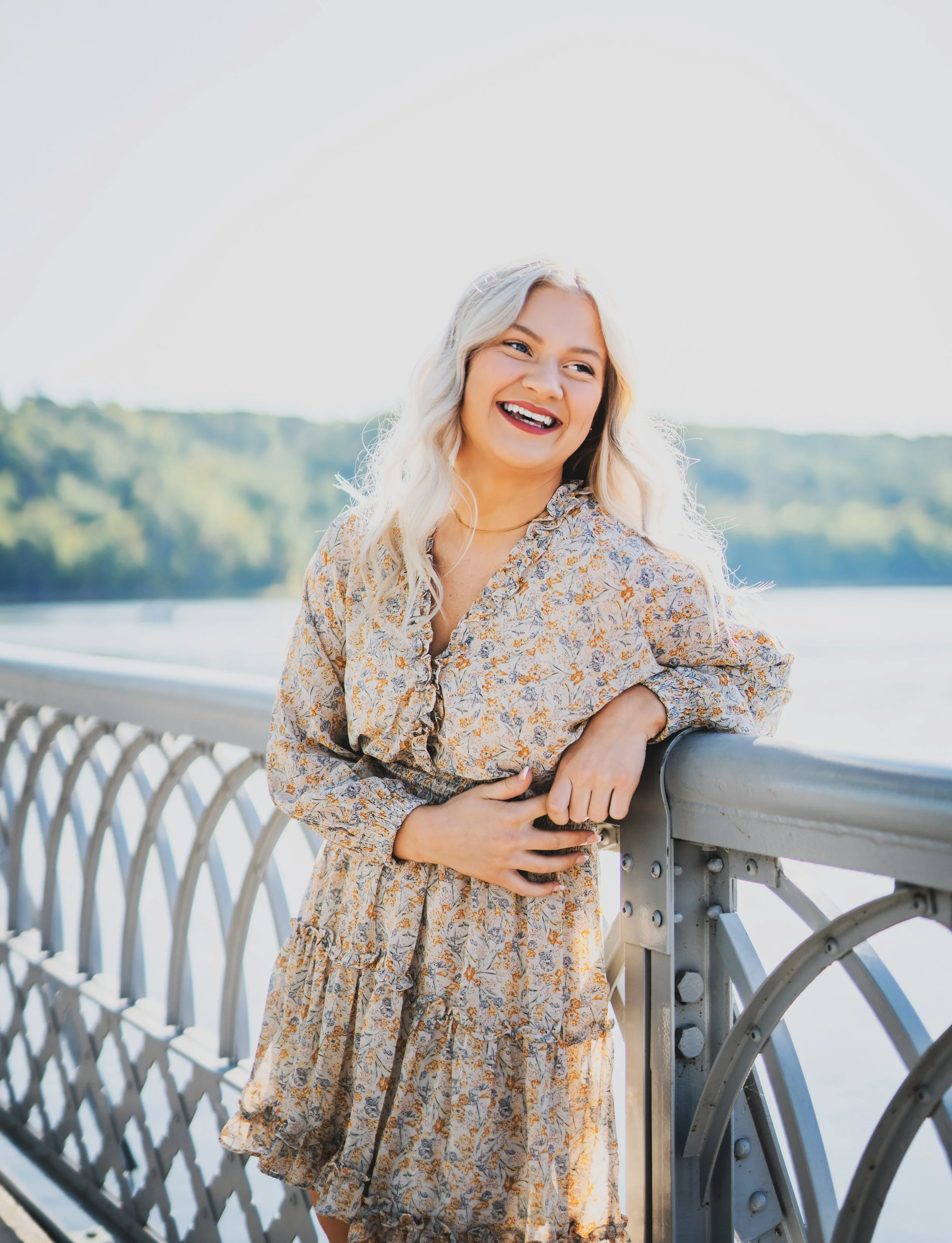 A young woman with blonde wavy hair smiling and laughing by a river, leaning on a metal railing with greenery in the background during daylight.