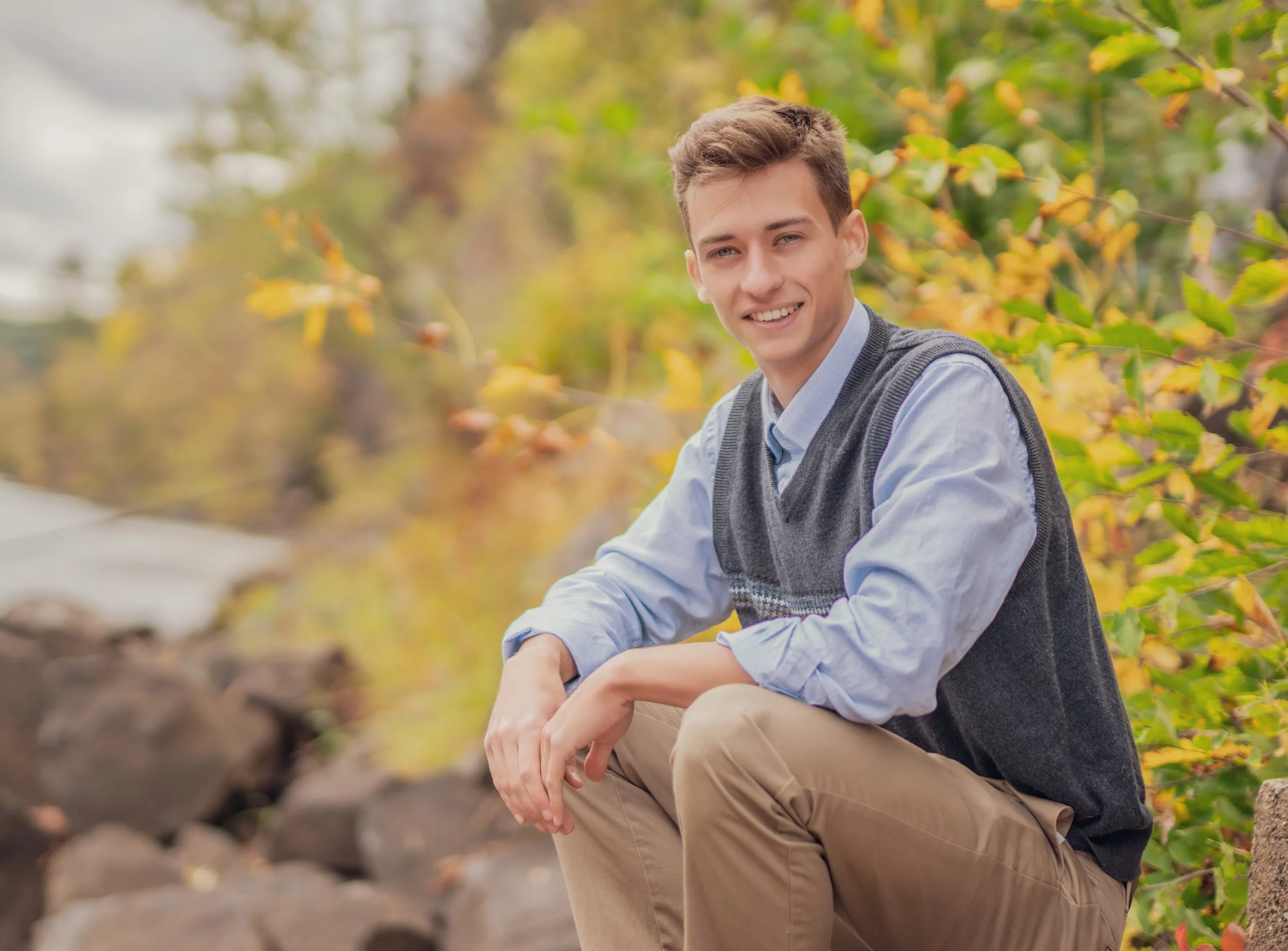 A young man sitting outdoors on rocks surrounded by autumn foliage, smiling at the camera.