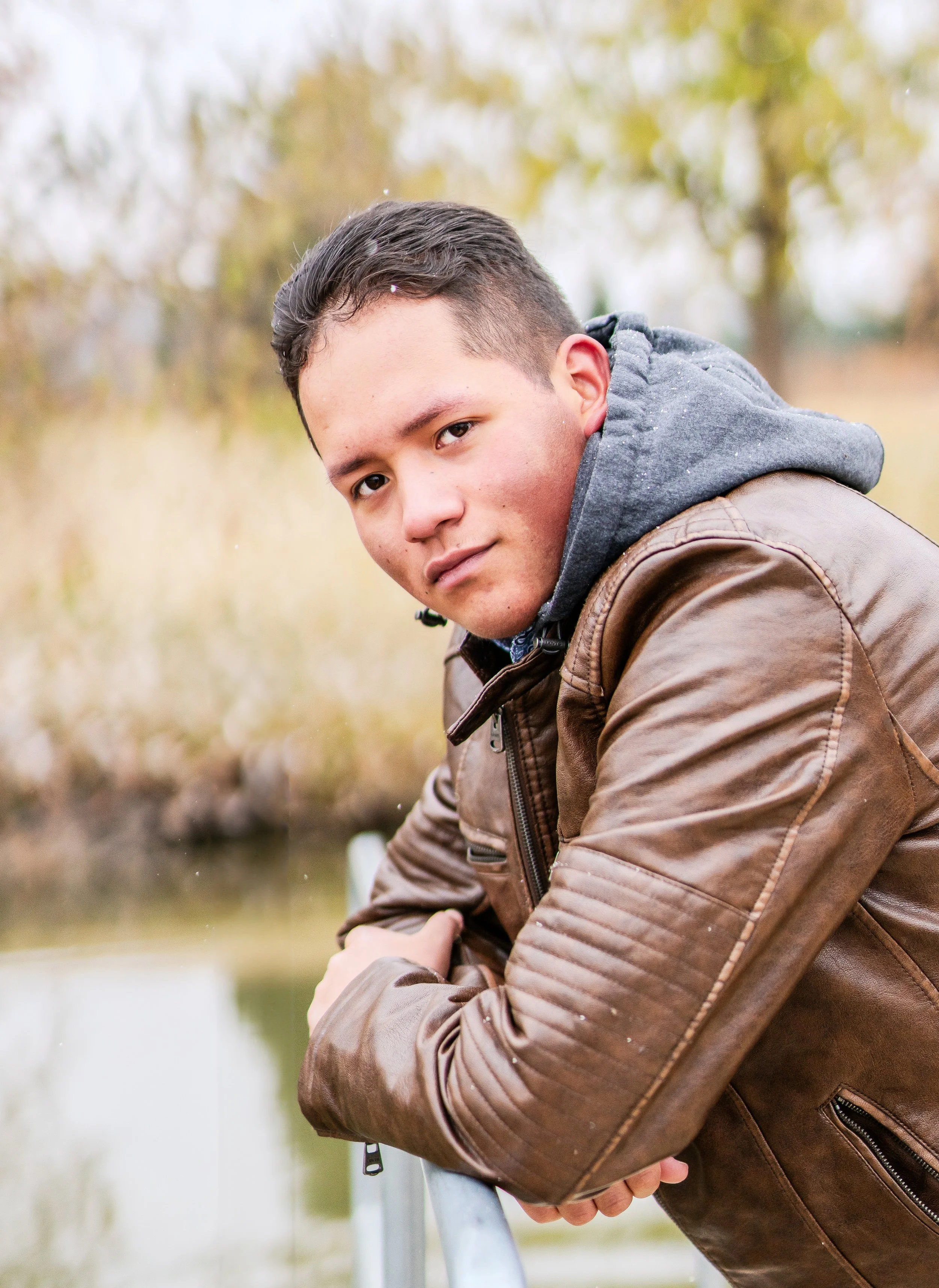 A young man leaning on a metal railing outdoors, wearing a brown leather jacket and a gray hoodie, with a body of water and autumn trees in the background.